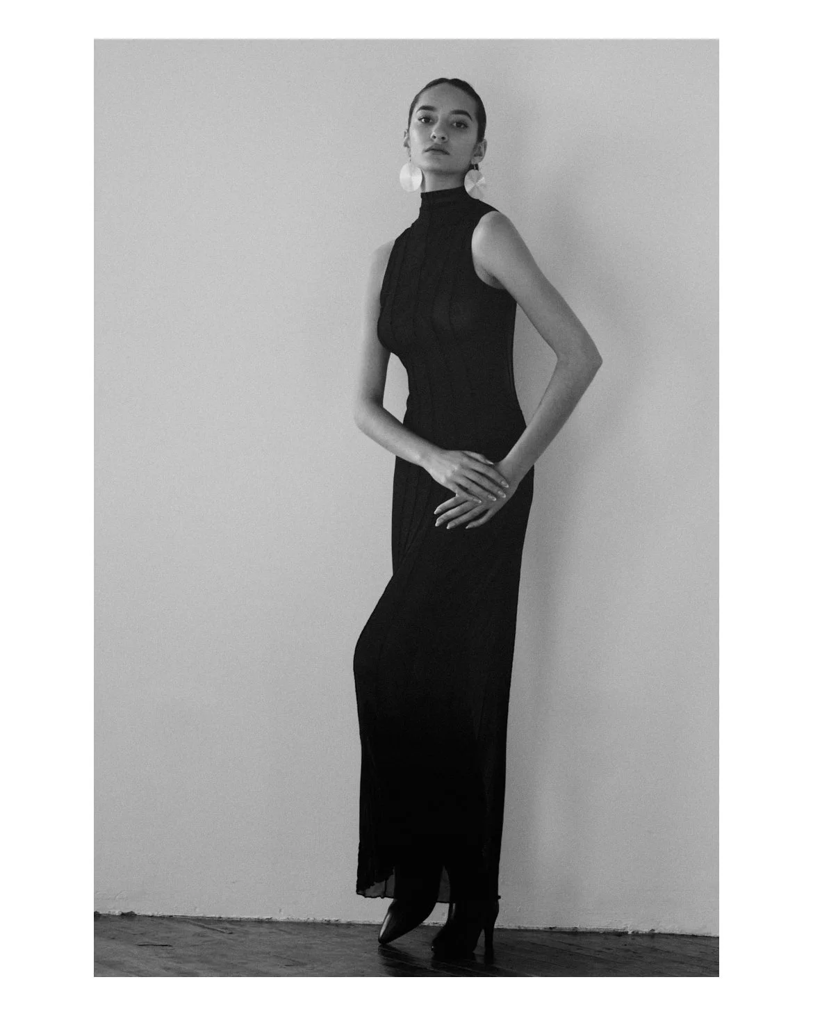 A woman in a black gown and high heels standing against a plain wall.