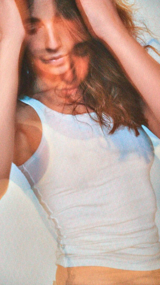 A woman is lying down with her eyes closed, smiling slightly, wearing a white tank top and holding her hair back with her right hand.