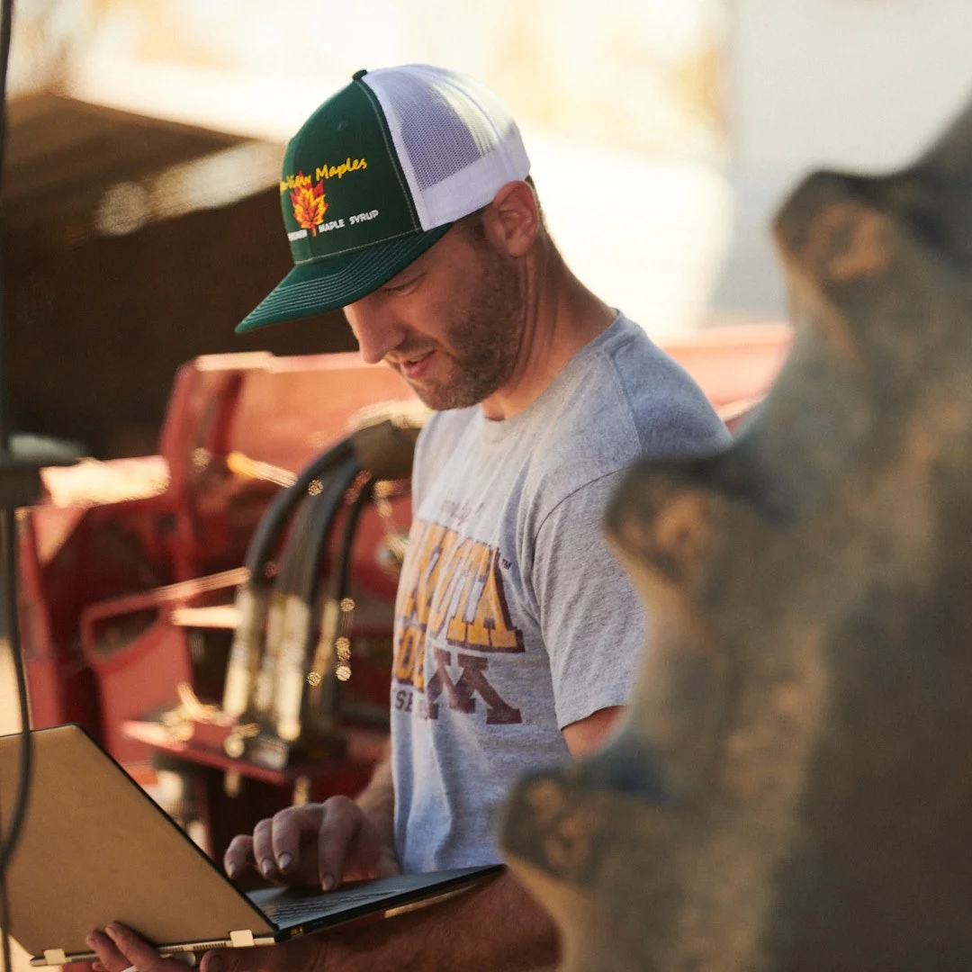 A bearded man wearing a baseball cap and a graphic T-shirt, looking down at a tablet or laptop, with a rustic background including a red tractor and a decorative bear sculpture.