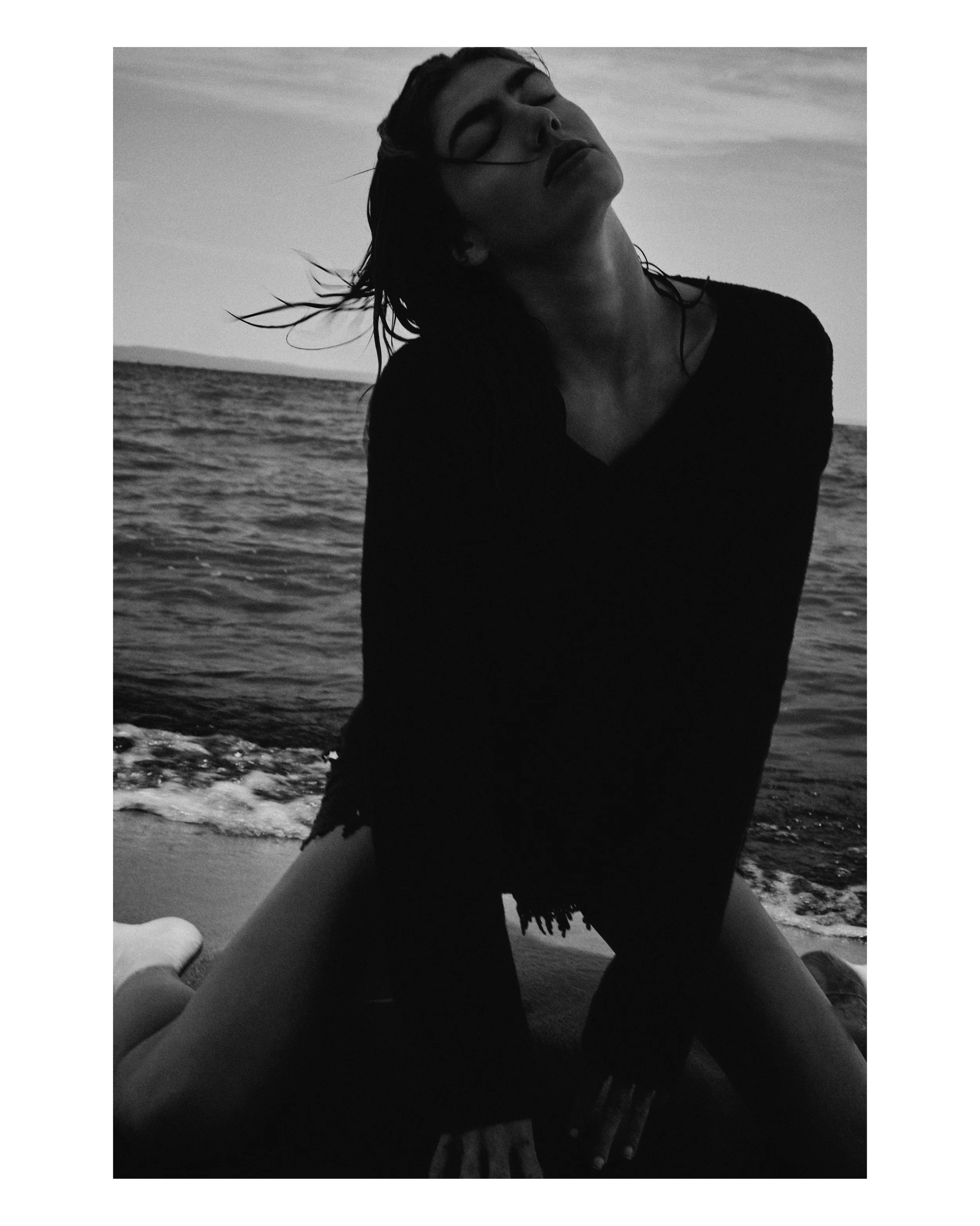 A black and white photo of a woman at the beach with her eyes closed, wet hair, wearing a long-sleeved top, kneeling on the sand with the ocean in the background.