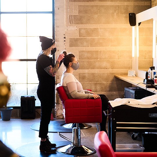 A woman sitting in a red salon chair gets her hair styled by a hairstylist in a salon with large windows and a beige brick wall.