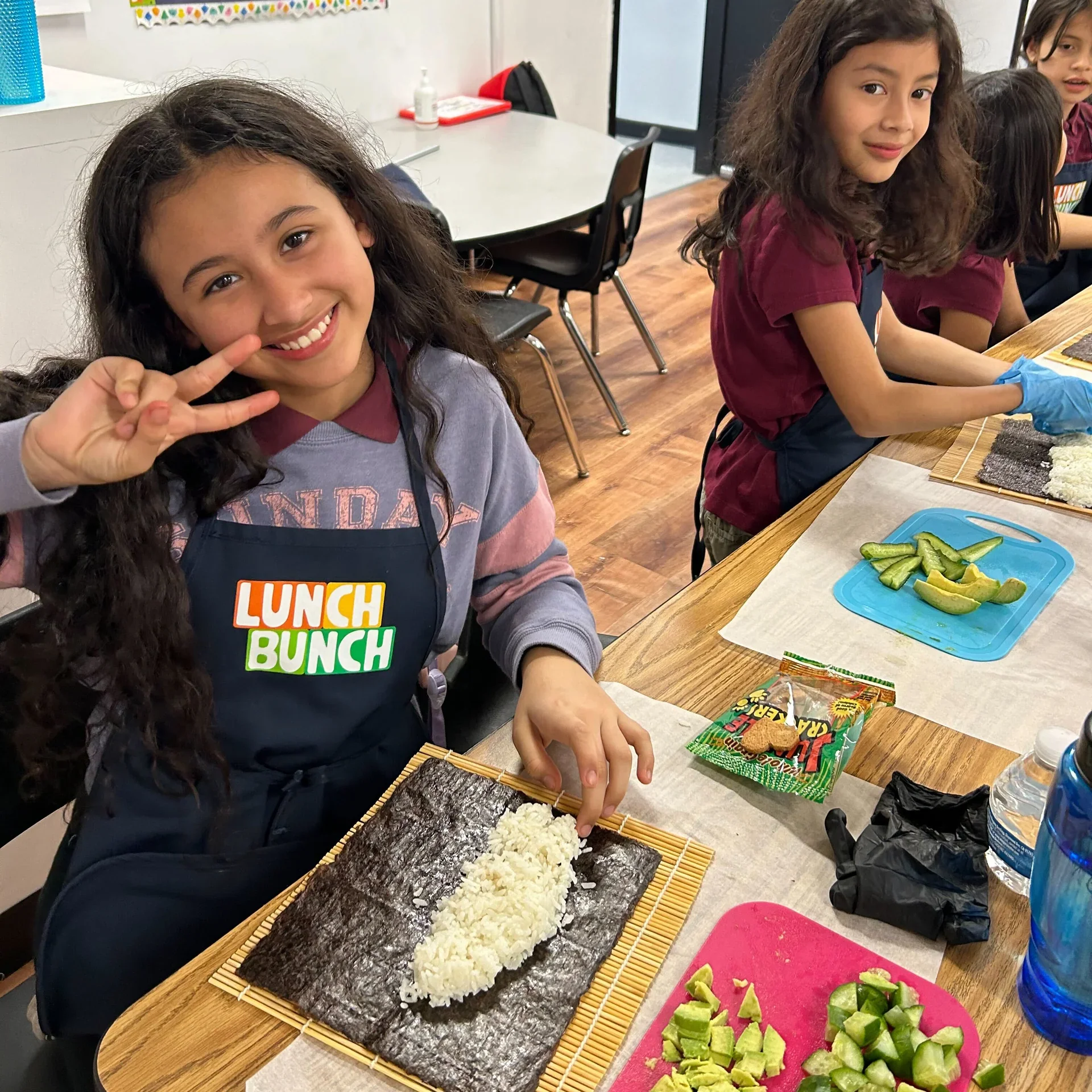 Two young girls preparing sushi in a classroom, with one girl smiling and the other focused on making sushi rolls. The table has ingredients including nori sheets, rice, cucumber, and avocado.