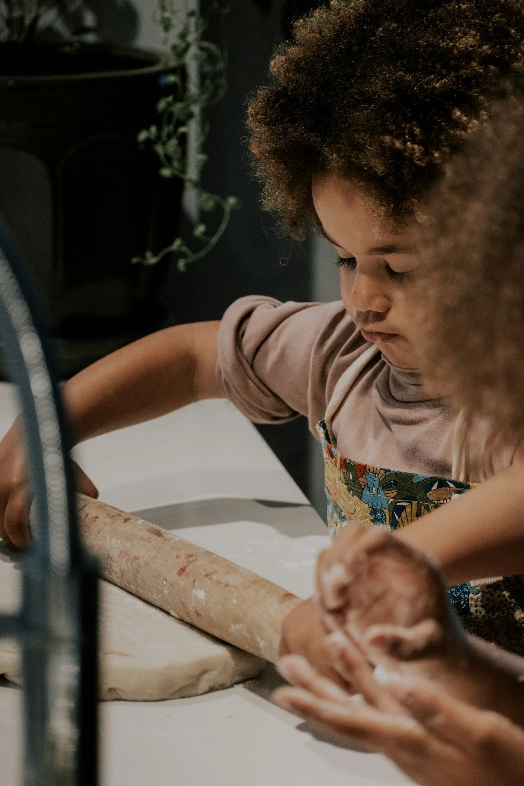 A young girl with curly hair rolling dough on a white surface, with flour scattered on it, in a kitchen setting.