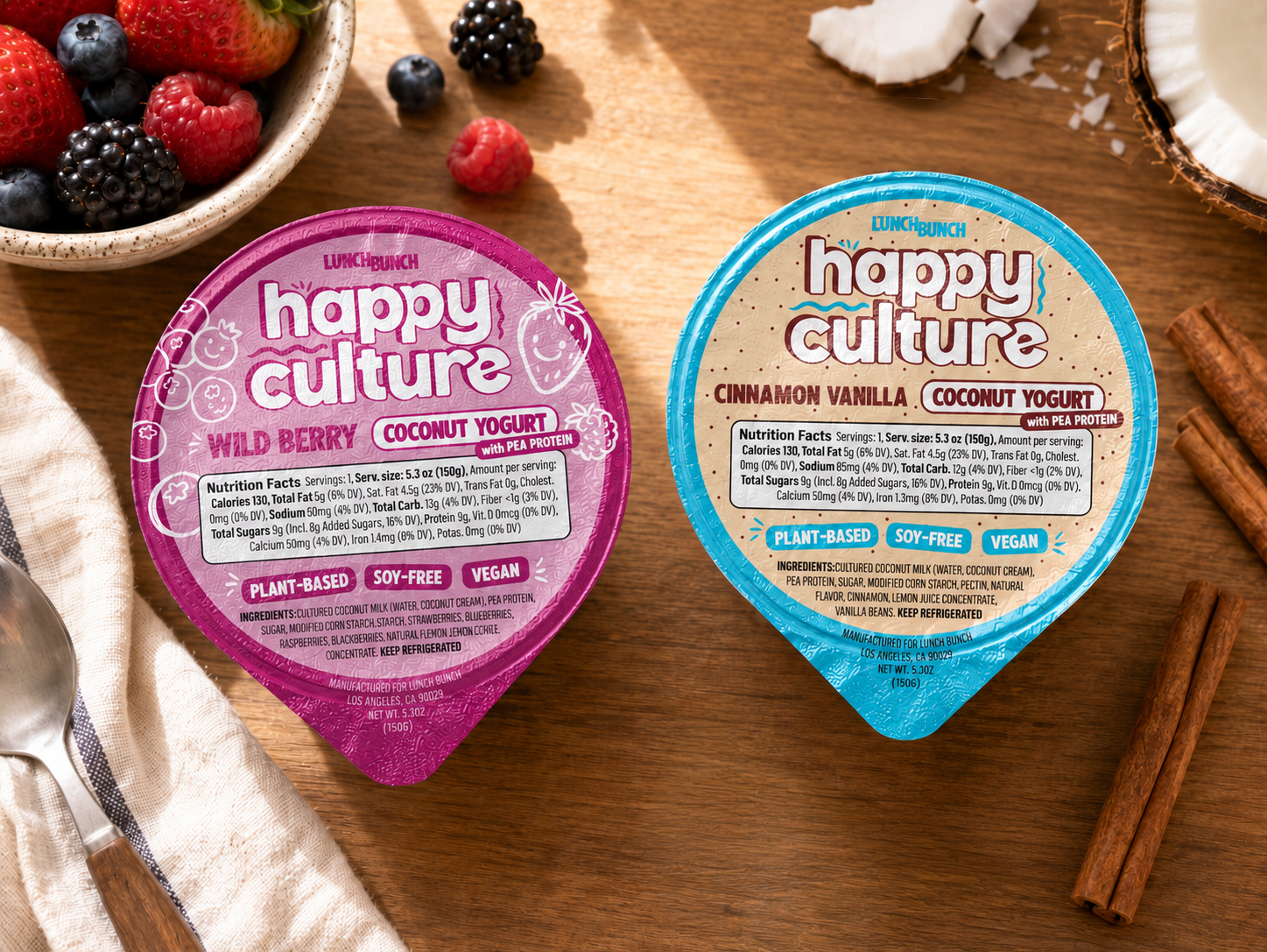Two containers of Lunch Bunch Happy Culture coconut yogurt on a wooden surface surrounded by fresh berries, sliced coconut, and cinnamon sticks.