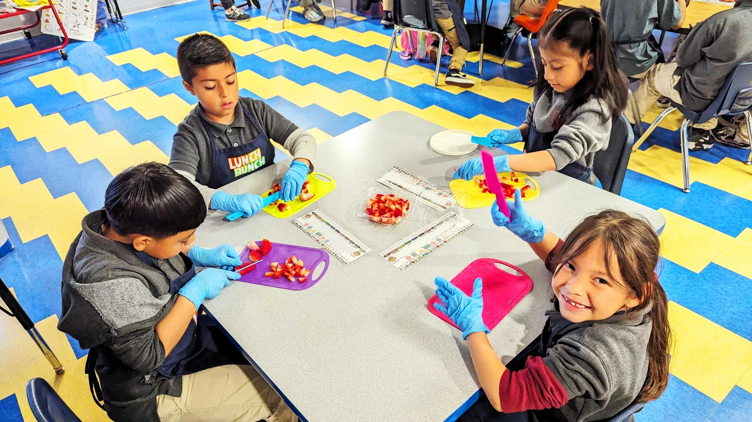 Children sitting at a table in a classroom, preparing to cut strawberries on colorful cutting boards, with some children smiling at the camera, wearing blue gloves.