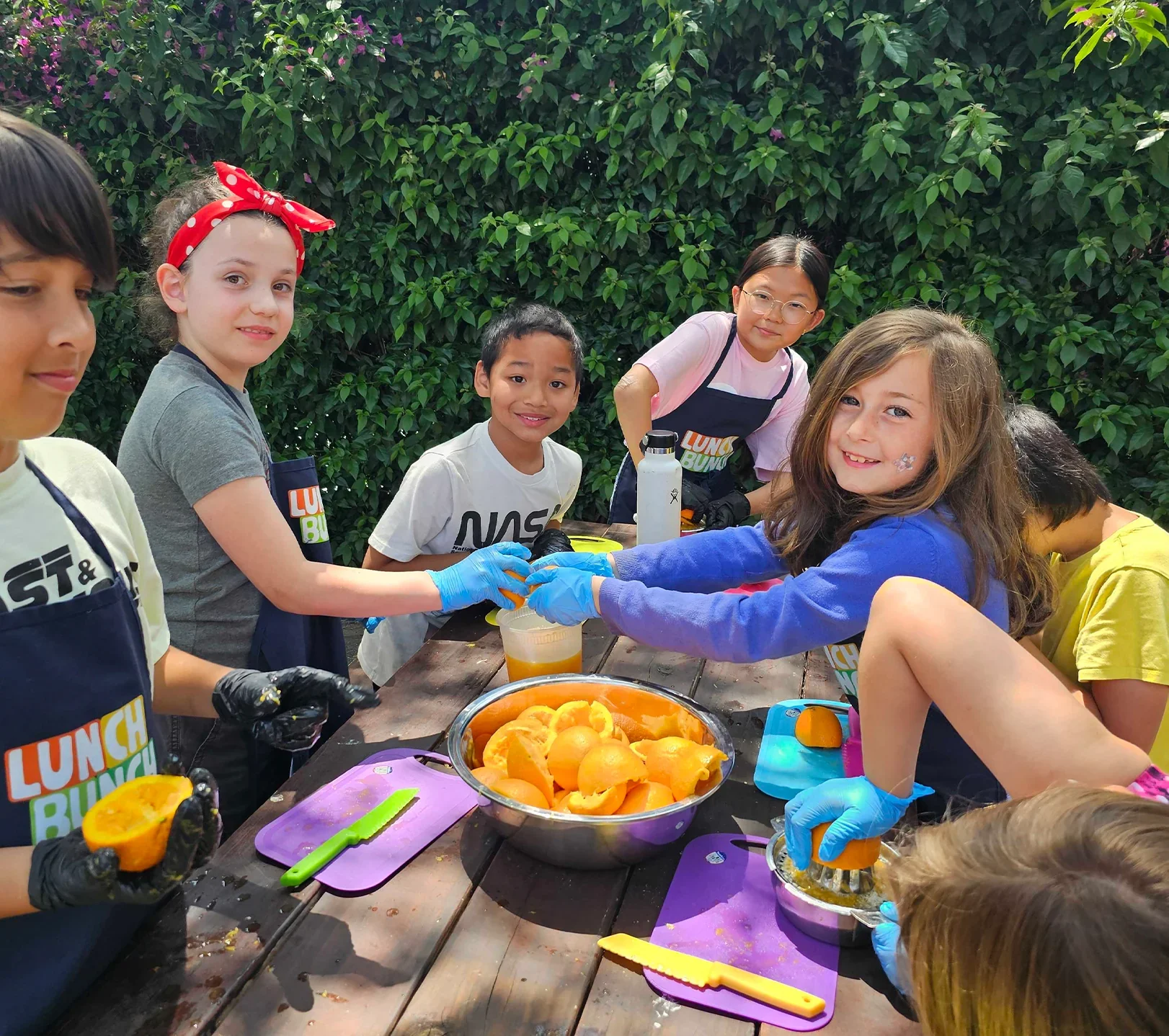 Children and a young woman gathered around a table outdoors, peeling oranges. They are wearing gloves and colorful aprons, with a large bowl of peeled oranges in the center. The background features lush greenery.