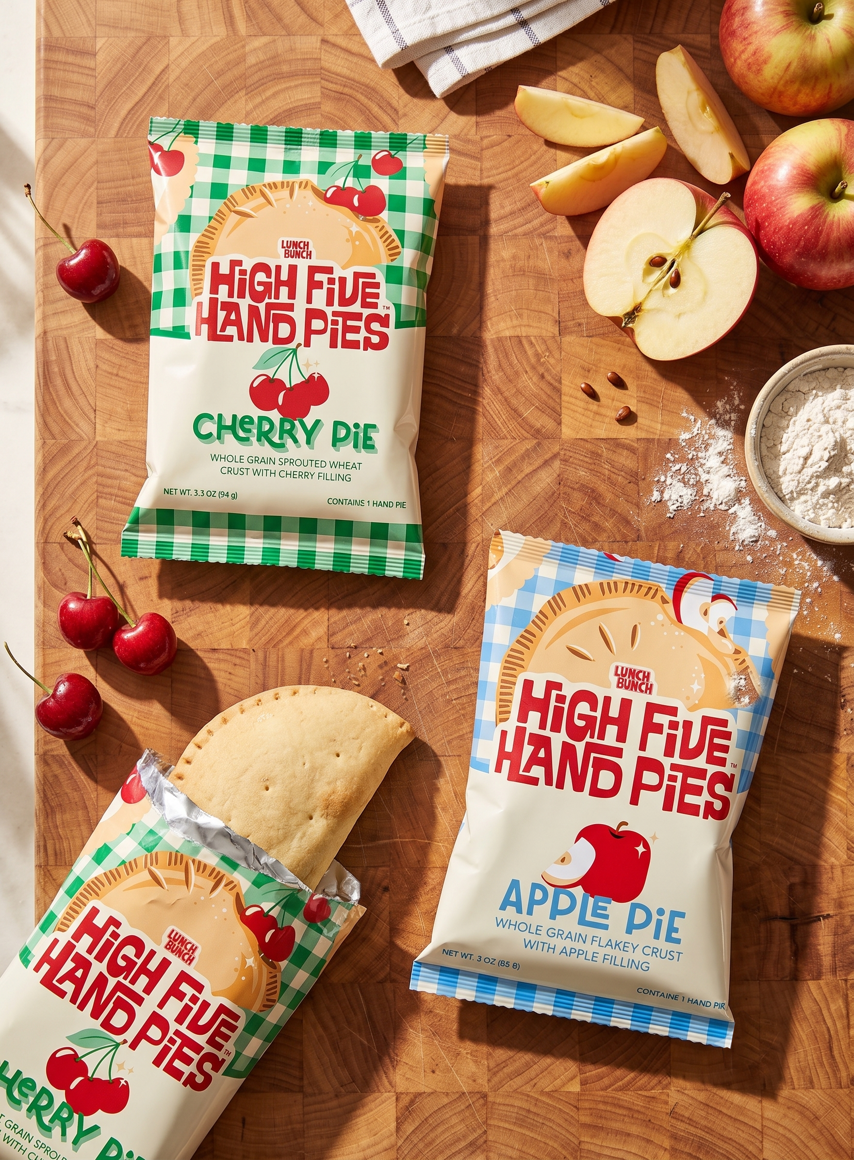 Two packages of High Five Hand Pies, Cherry Pie and Apple Pie flavor, on a wooden surface with fresh cherries, apple slices, and a bowl of flour nearby.