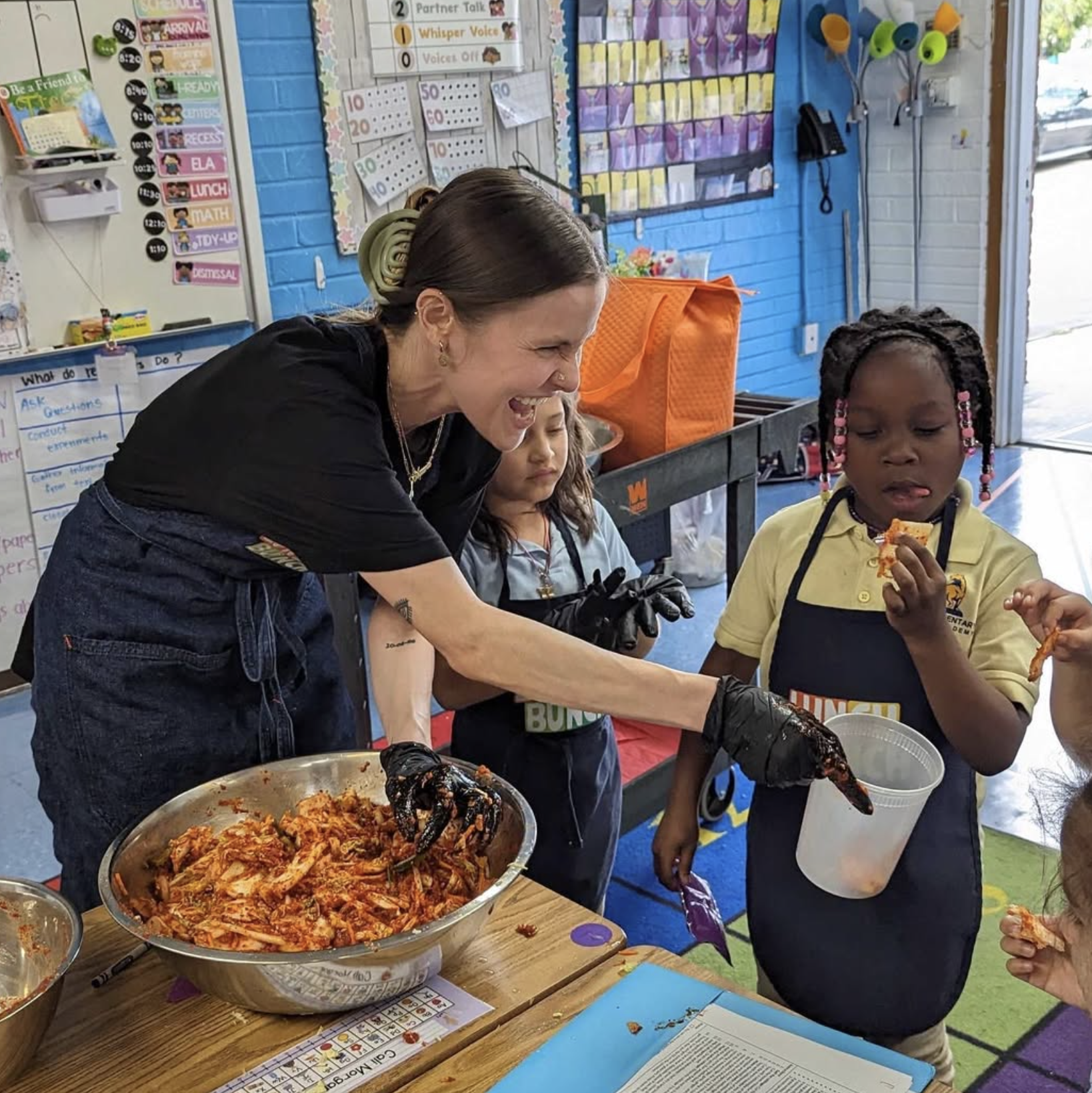 A classroom setting where a woman is serving food, specifically a large bowl of pasta with sauce, to children. The children are wearing school uniforms, and one girl is holding a container and eating. The background shows colorful educational posters, a blue wall, and classroom supplies.