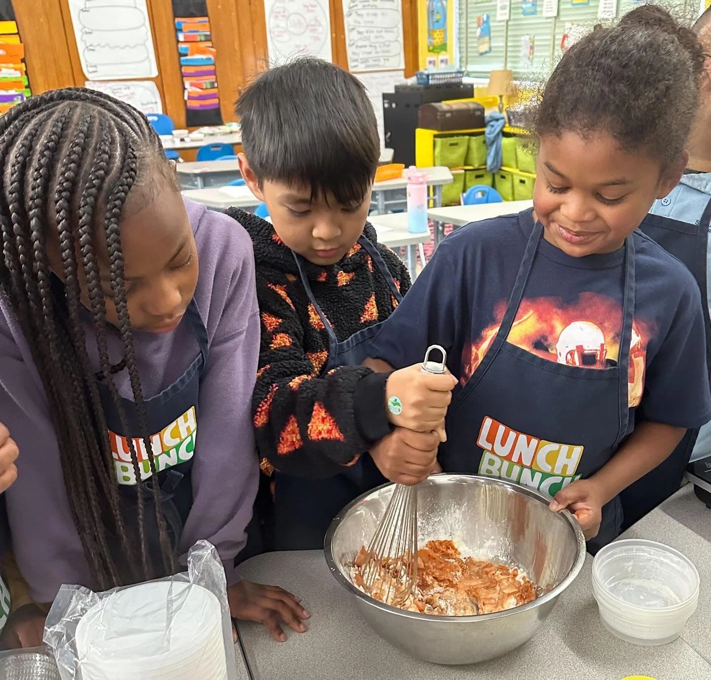 Three children wearing aprons are mixing ingredients in a large stainless steel bowl in a classroom setting.