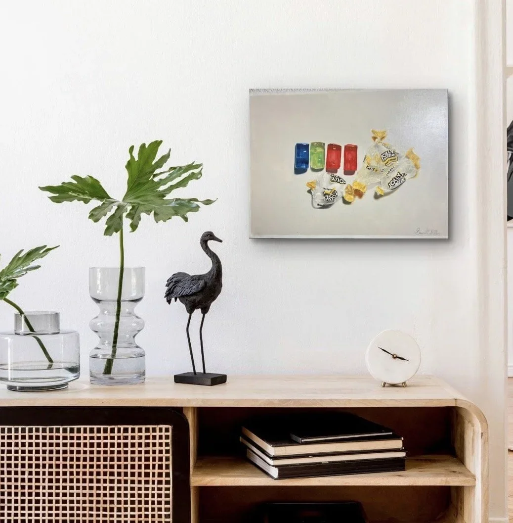A modern interior decor scene featuring a wooden console table with books, a decorative black crane sculpture, two glass vases with large green leaves, a white round clock, and a white painting with colorful candy wrappers on the wall.