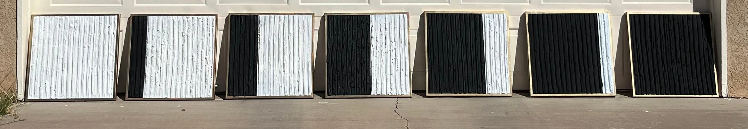 Six framed abstract artworks with textured vertical lines, arranged in a row outdoors against a wall, alternating in black and white colors.