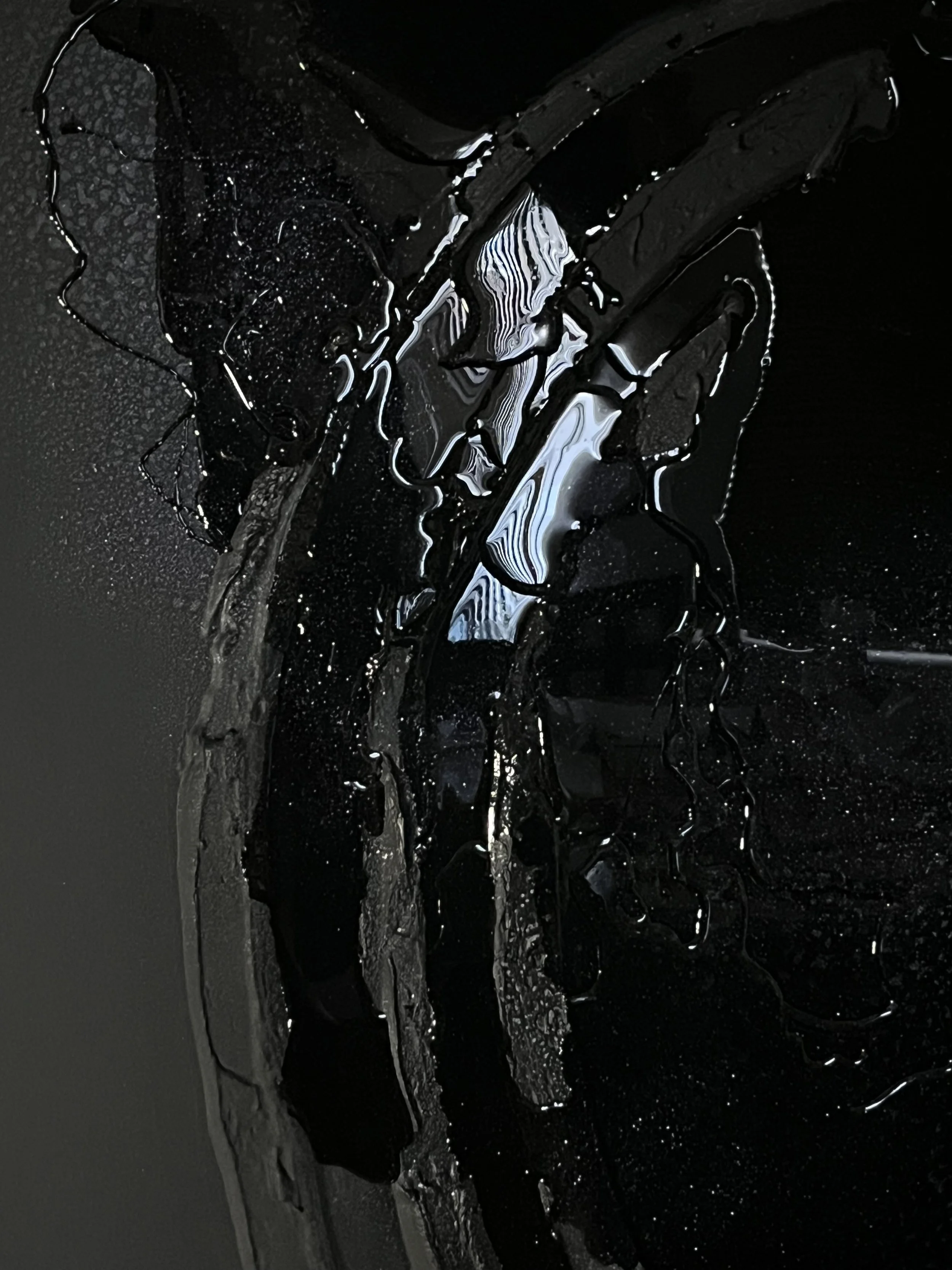 Close-up photograph of melting black wax with drips and cracks, having a shiny, glossy texture on a dark background.