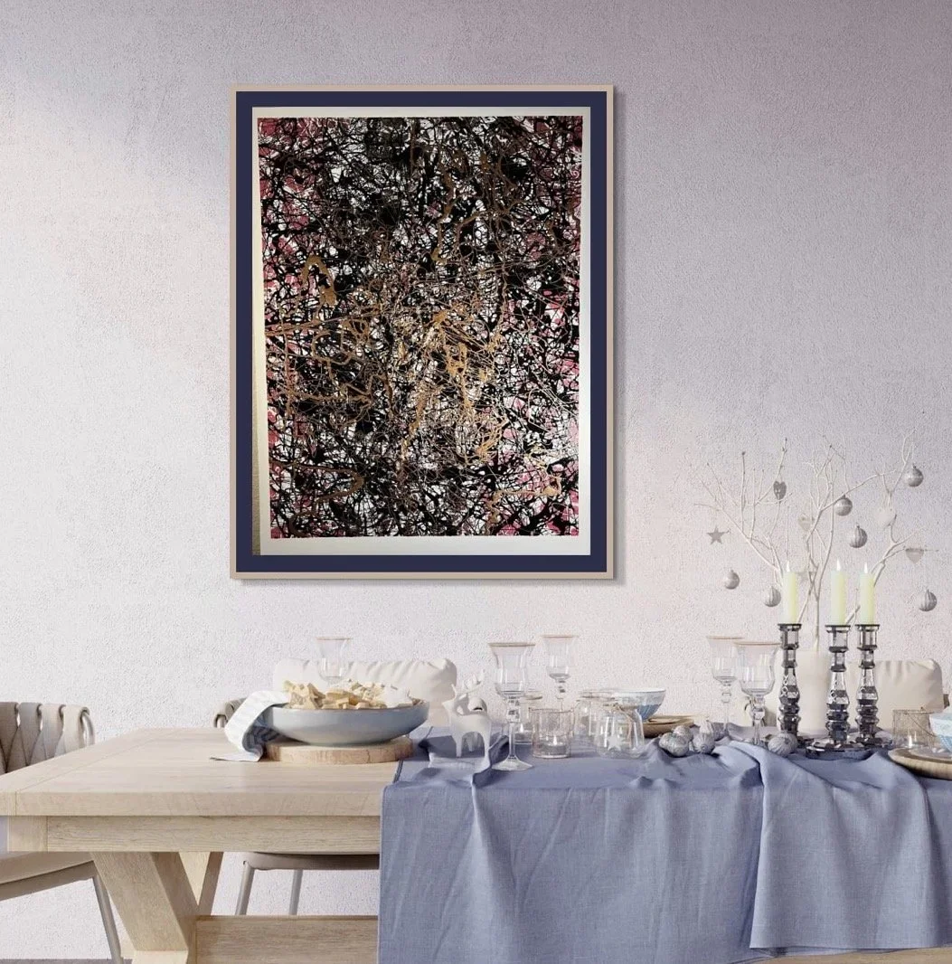 A dining room with a wooden table covered with a blue tablecloth, decorated with glassware, candles, and silver ornaments. An abstract artwork with a black, brown, and pink pattern hangs on the wall above.