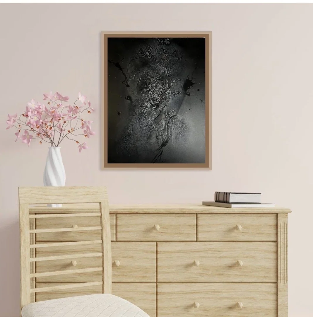 A wooden dresser with a stack of books on top, a white vase with pink flowers, and a framed abstract black and gray artwork on a light-colored wall.