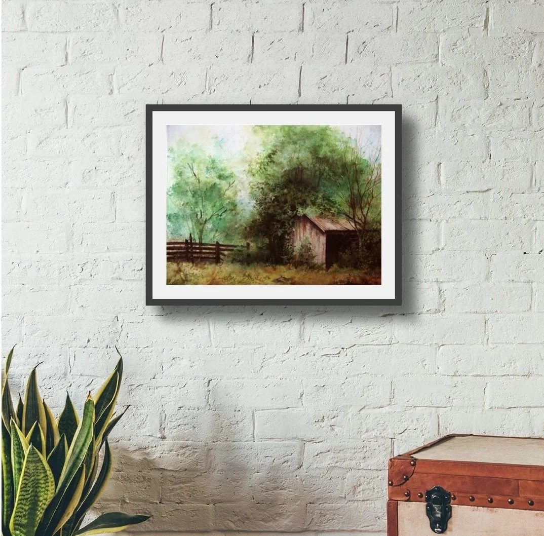A framed watercolor painting of a rustic barn surrounded by green trees hangs on a white brick wall. A potted plant with long green and yellow striped leaves is in the lower left corner, and a wooden chest with metal hardware is in the lower right corner.