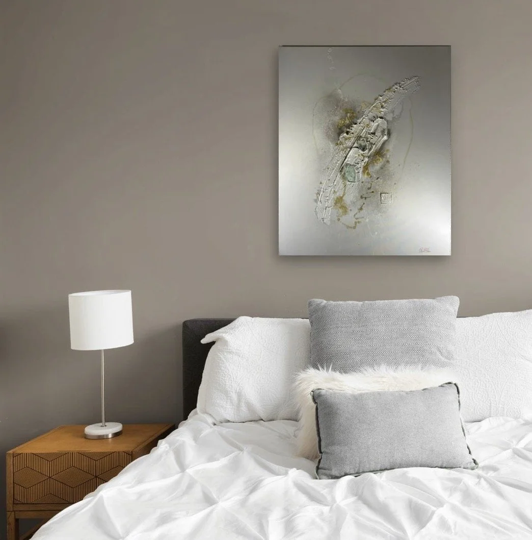 Modern bedroom with gray wall, abstract art painting above bed, white bedding, gray and fur pillows, wooden nightstand with white lamp.