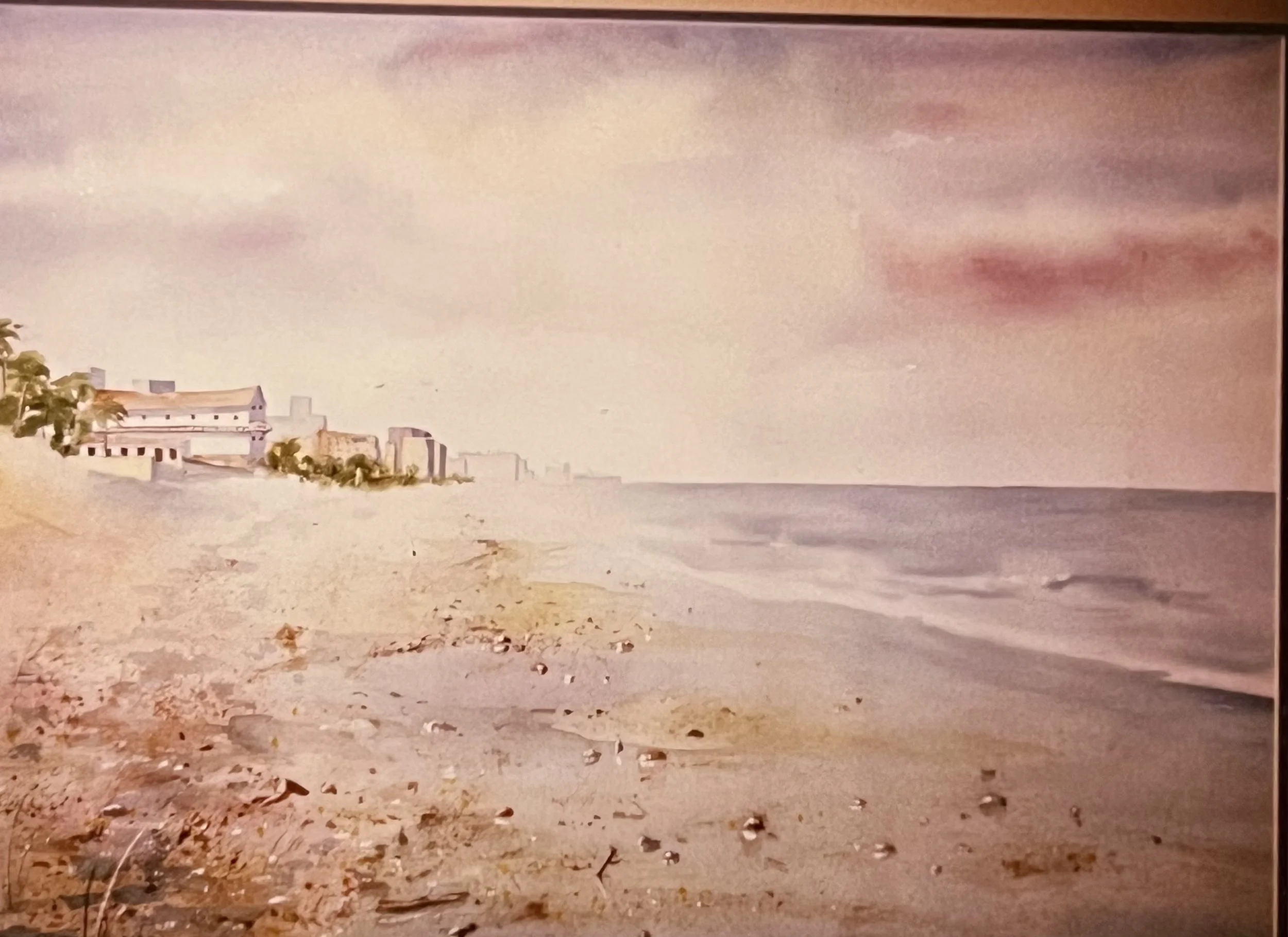A beach scene with a sandy shore, calm water, and overcast sky. In the background, there are buildings, including a castle and other structures, along the coastline.