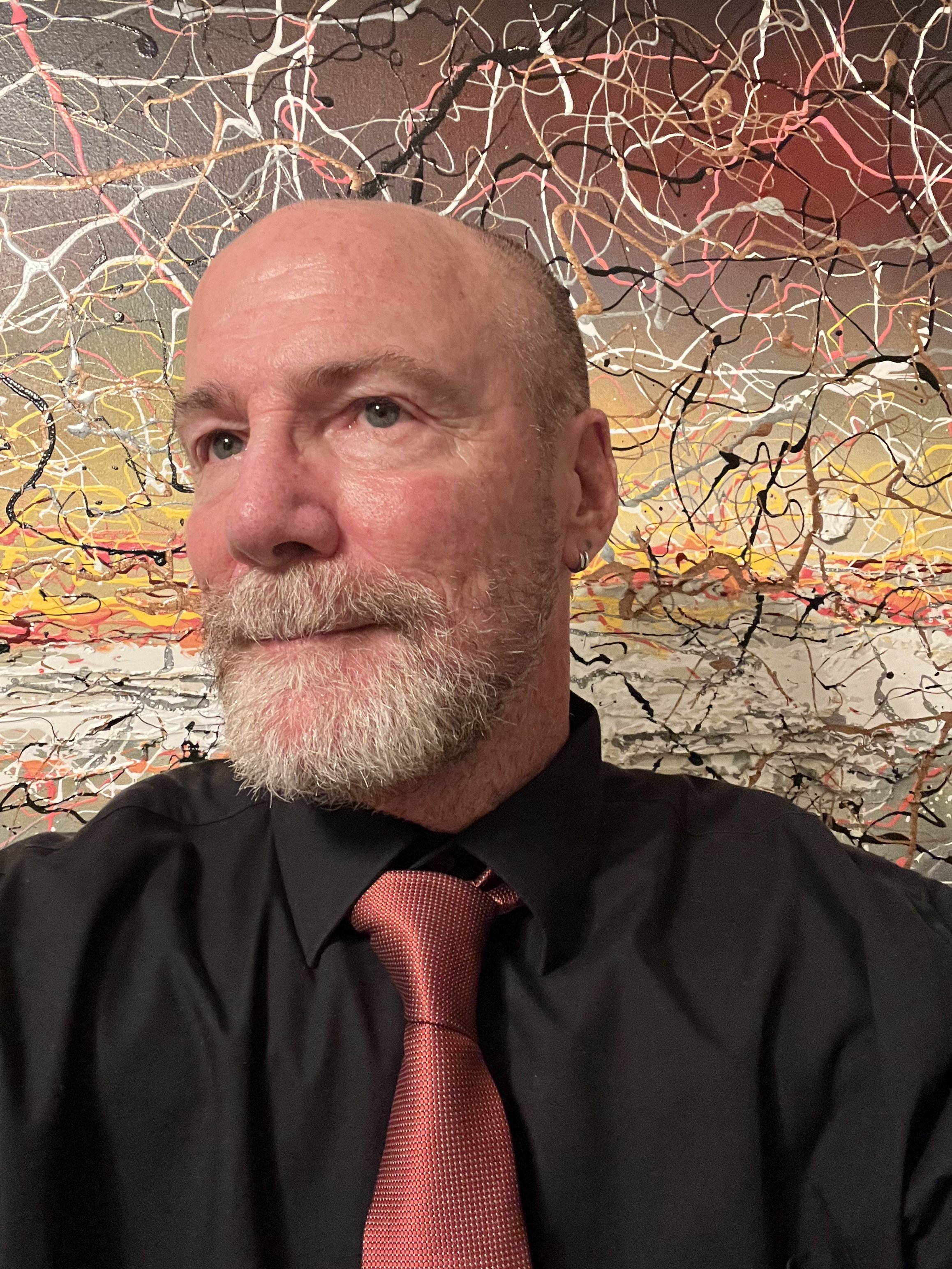 A close-up of a middle-aged man with a bald head and a gray beard, wearing a black shirt and a red tie. Behind him, there is an abstract art piece with colorful intertwining lines on a textured wall.
