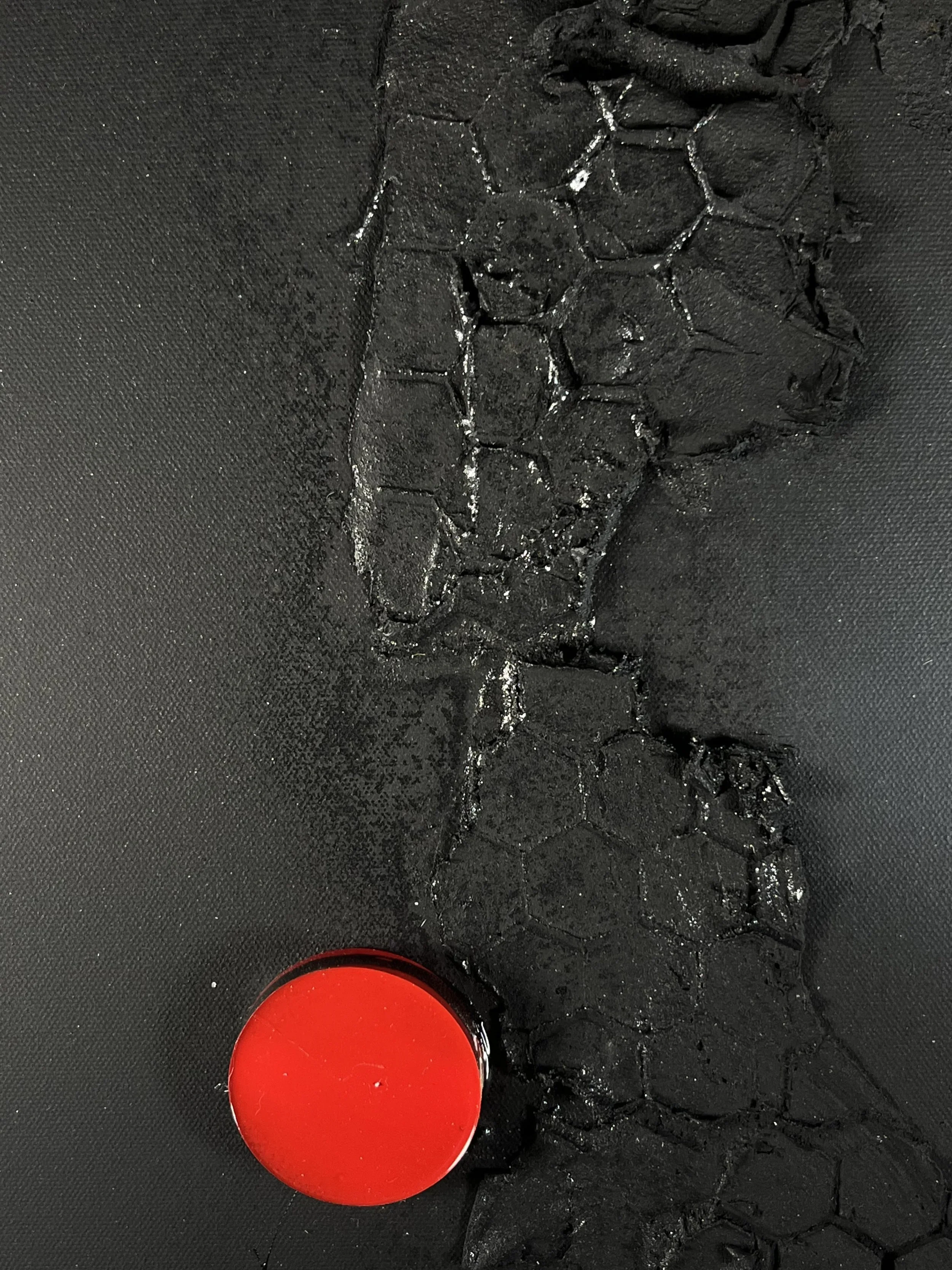 A large red button on a textured black surface. The button appears to be made of shiny plastic.
