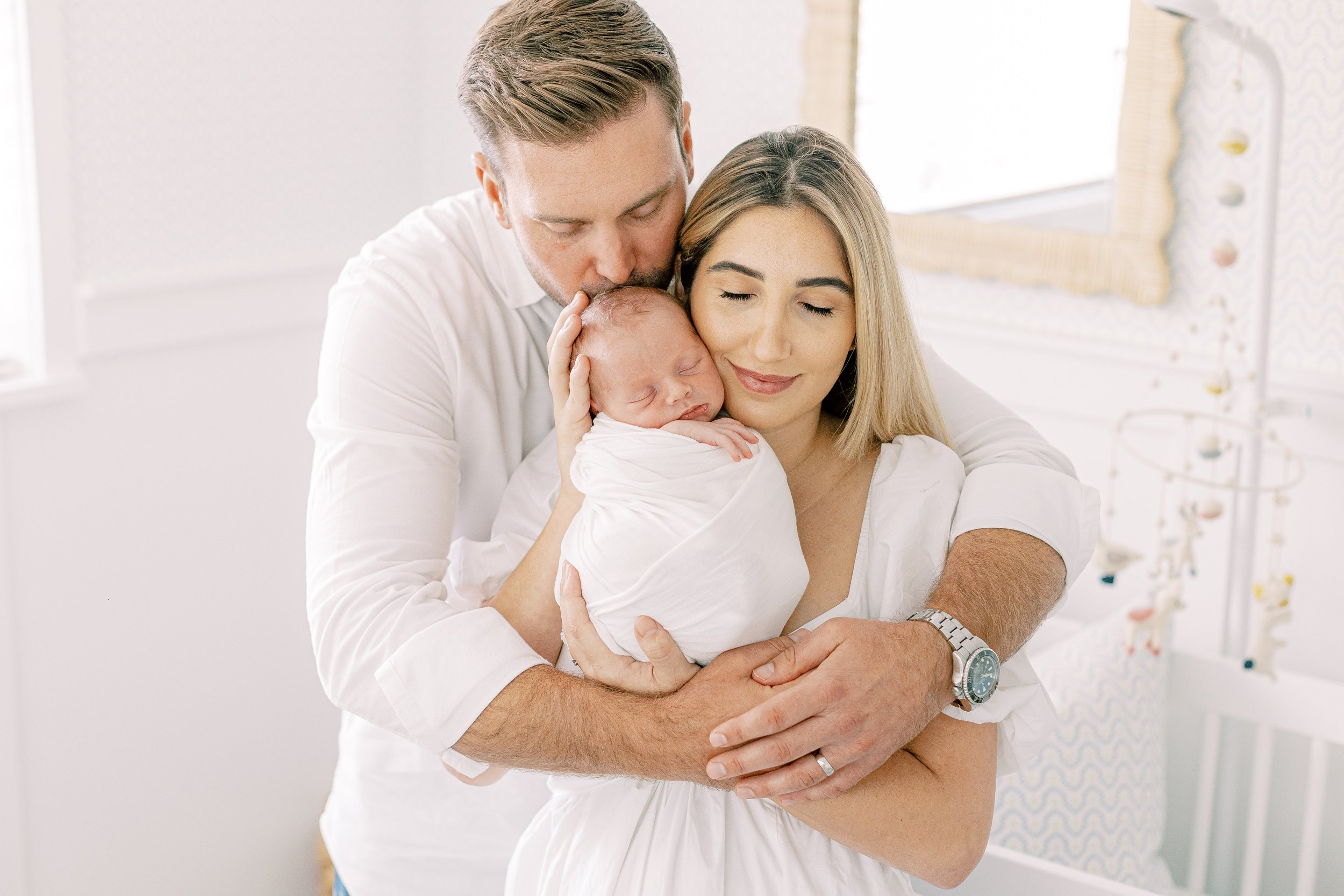 A man and woman in a nursery holding and cuddling a newborn baby, with the man kissing the baby and the woman smiling with eyes closed.