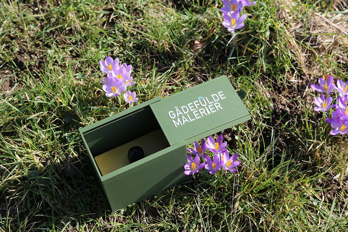 En grøn flyttekasse med teksten "GÅDEFØLDE MALERIER" står på græs omgivet af lilla krokus blomster.