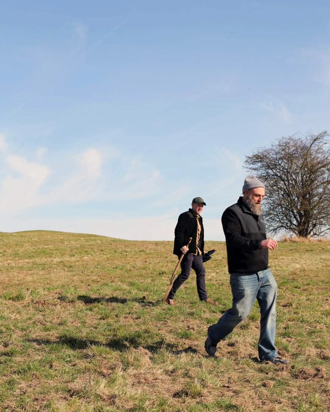 To mænd går gennem en åben græsmark under en klar blå himmel. Den foran mand har gråt hår og skæg, og den bagved mand har cap og holder en stok.