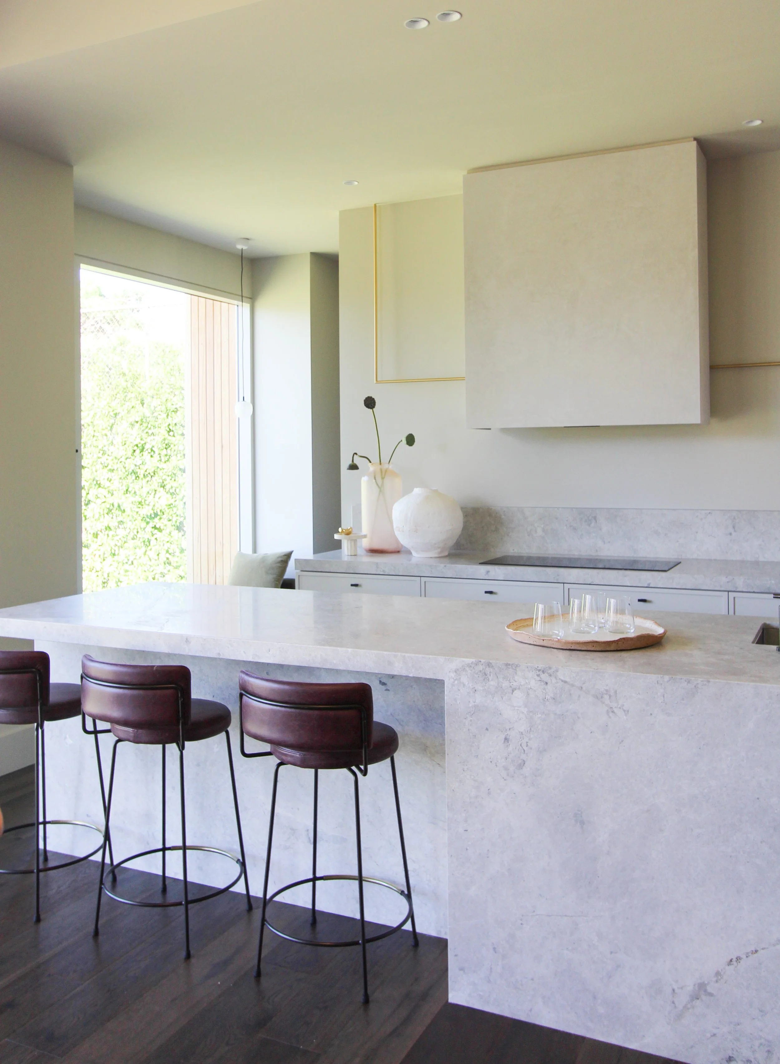 Modern kitchen with a large white marble island, three brown bar stools, a window letting in sunlight, decorative vases on the counter, and minimalistic design.
