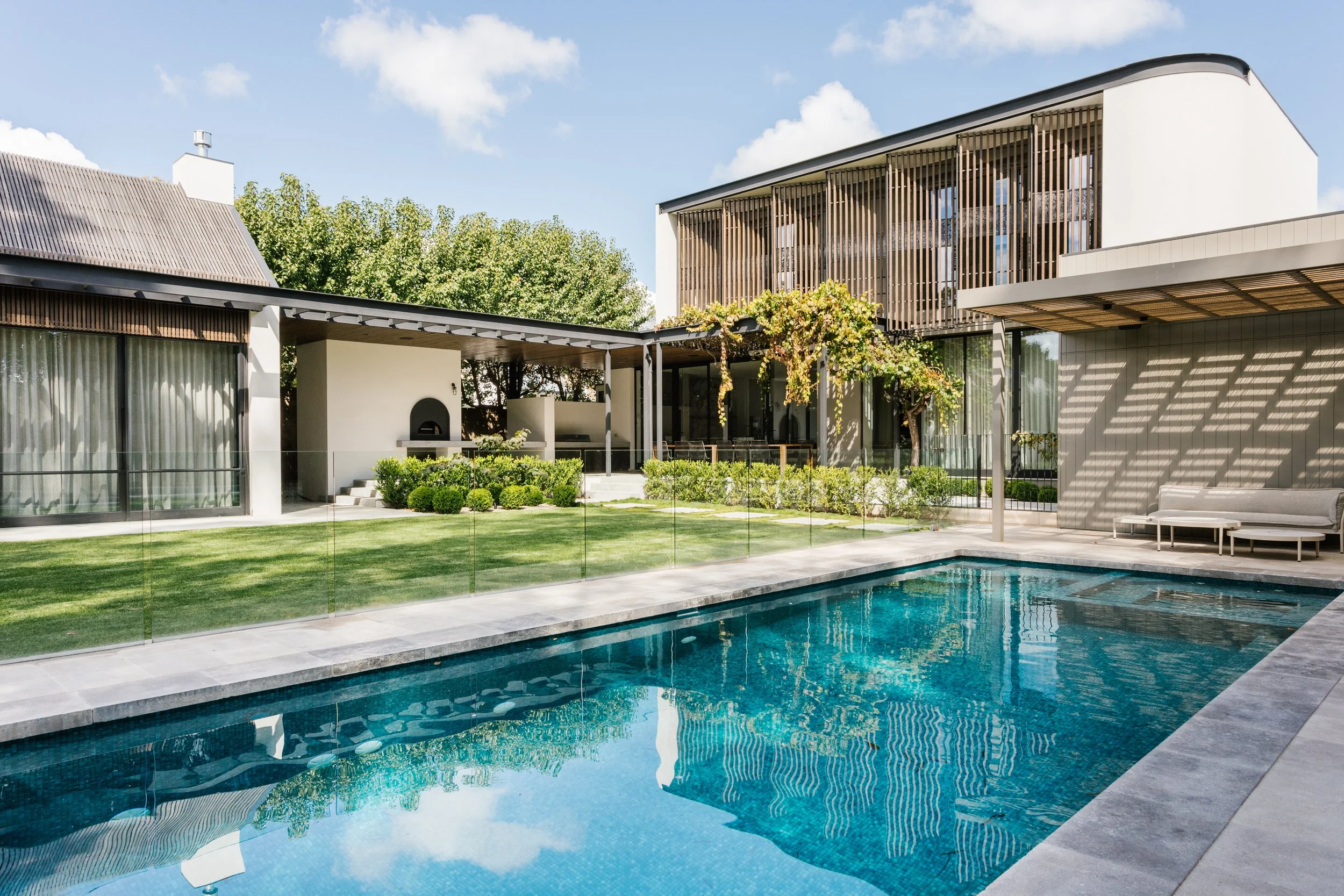 Modern house with a swimming pool, manicured lawn, and outdoor seating area, with a lush green tree in the background.