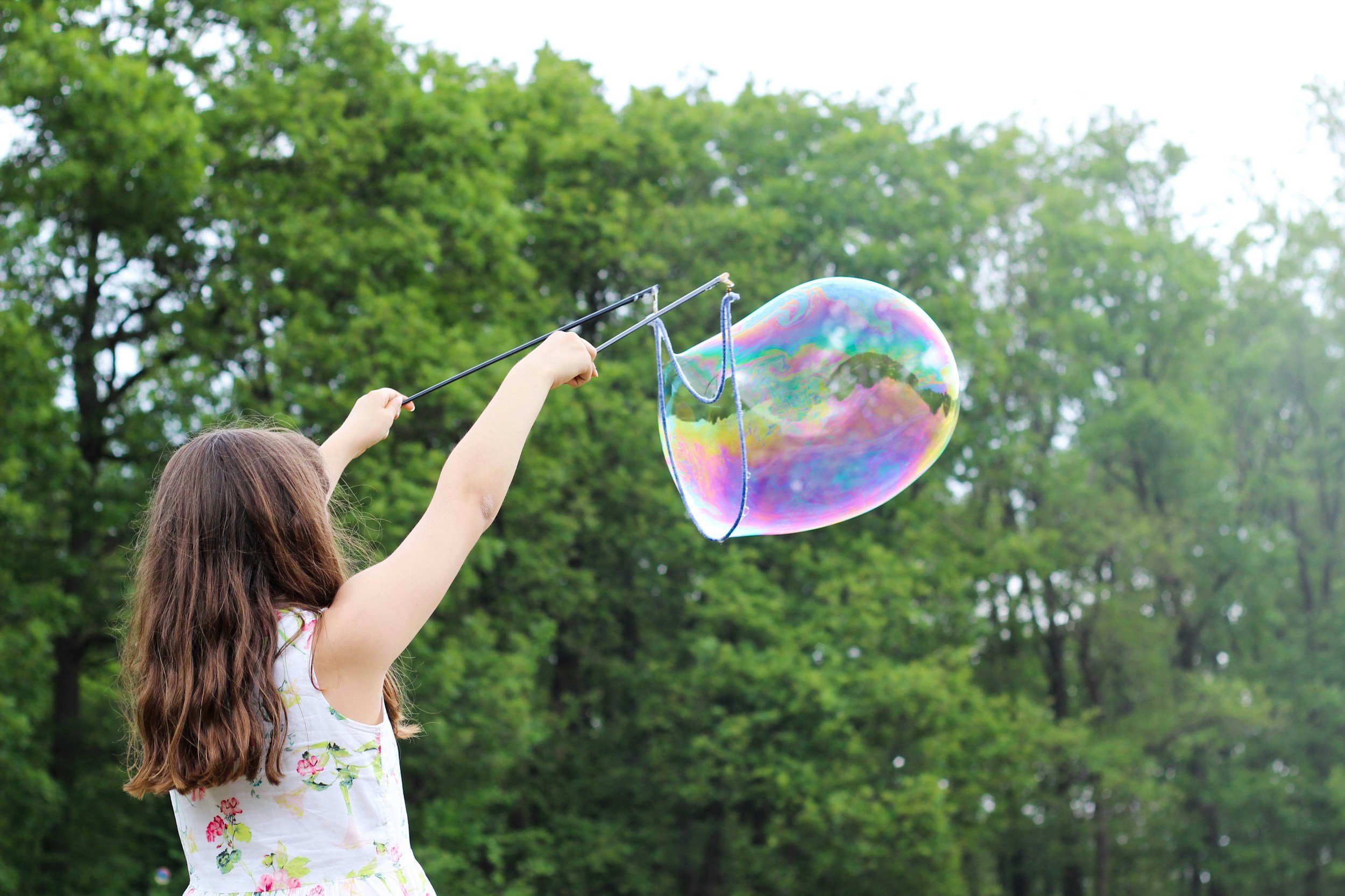 A young girl holding a bubble wand with a large colorful soap bubble in a park surrounded by green trees.