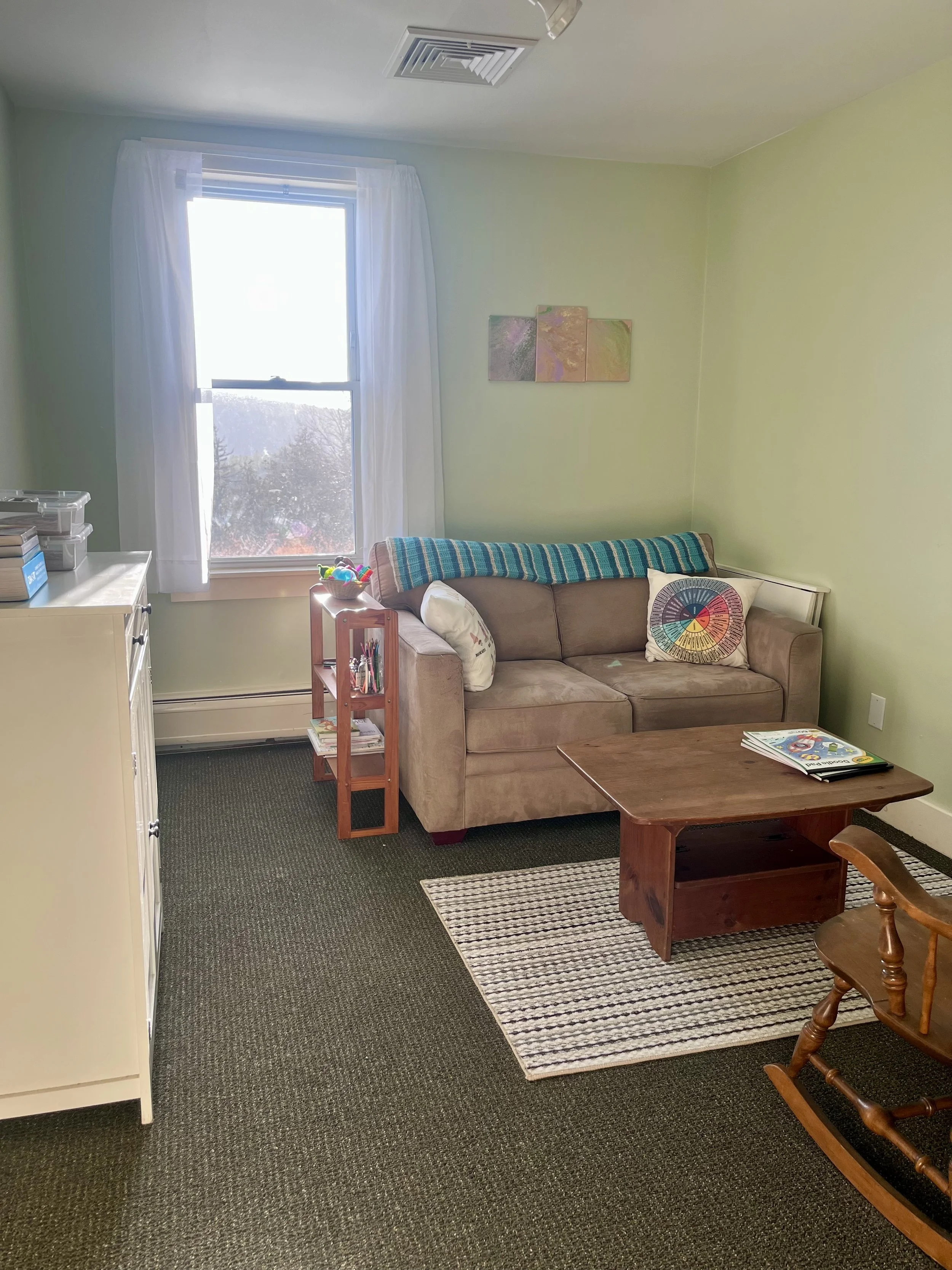 Office with a beige couch, wooden coffee table, rocking chair, window with white curtains, small shelf with books and toys, and wall art on a light green wall.