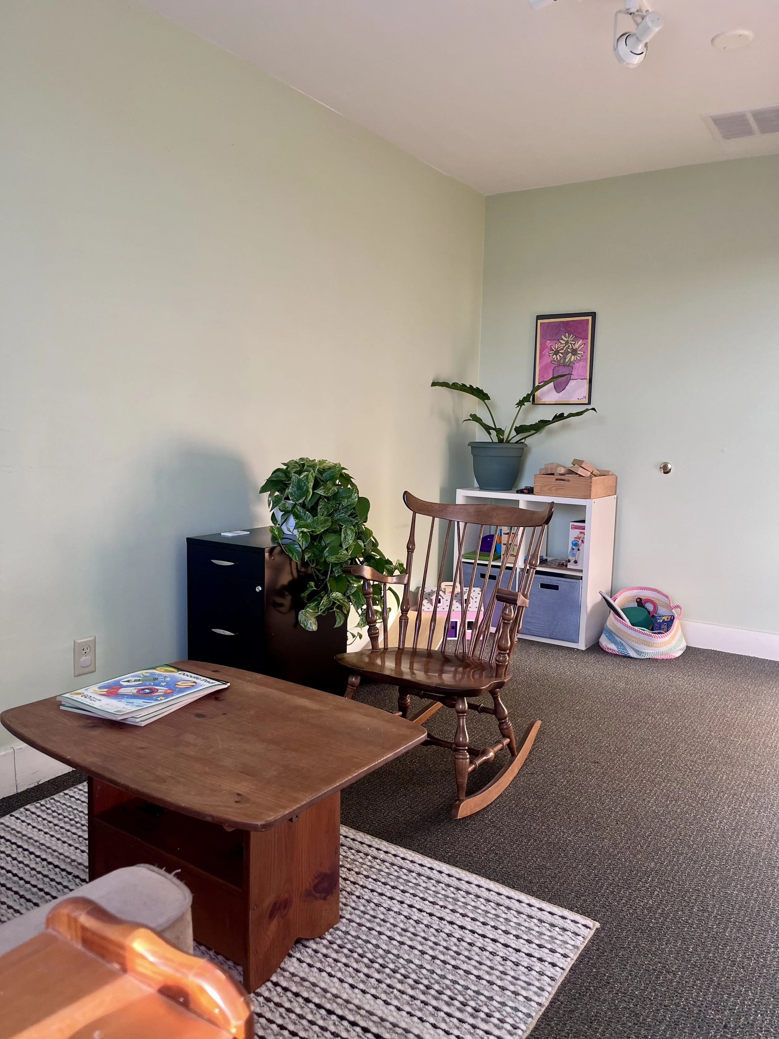 A cozy office with light green walls and a gray carpet, featuring a wooden rocking chair, a small black cabinet with a potted plant, a bookshelf with toys, a colorful basket, and a framed artwork of flowers on the wall.