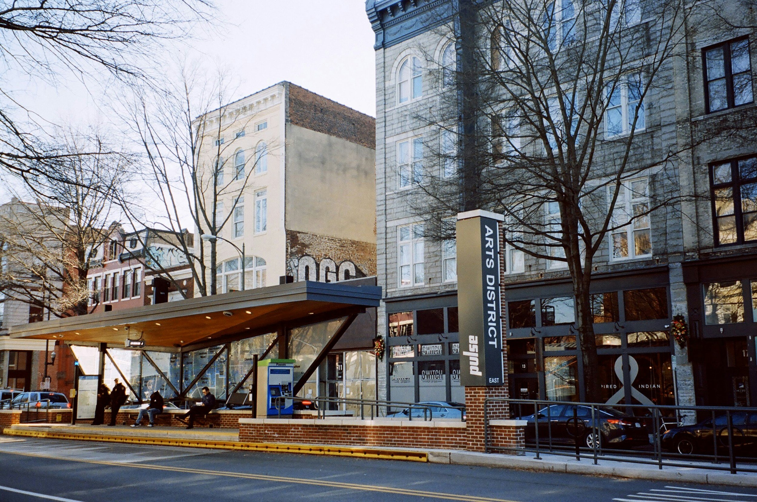 Bus stop on a city street with benches, people sitting and standing, a sign reading 'Arts District' and buildings with leafless trees.