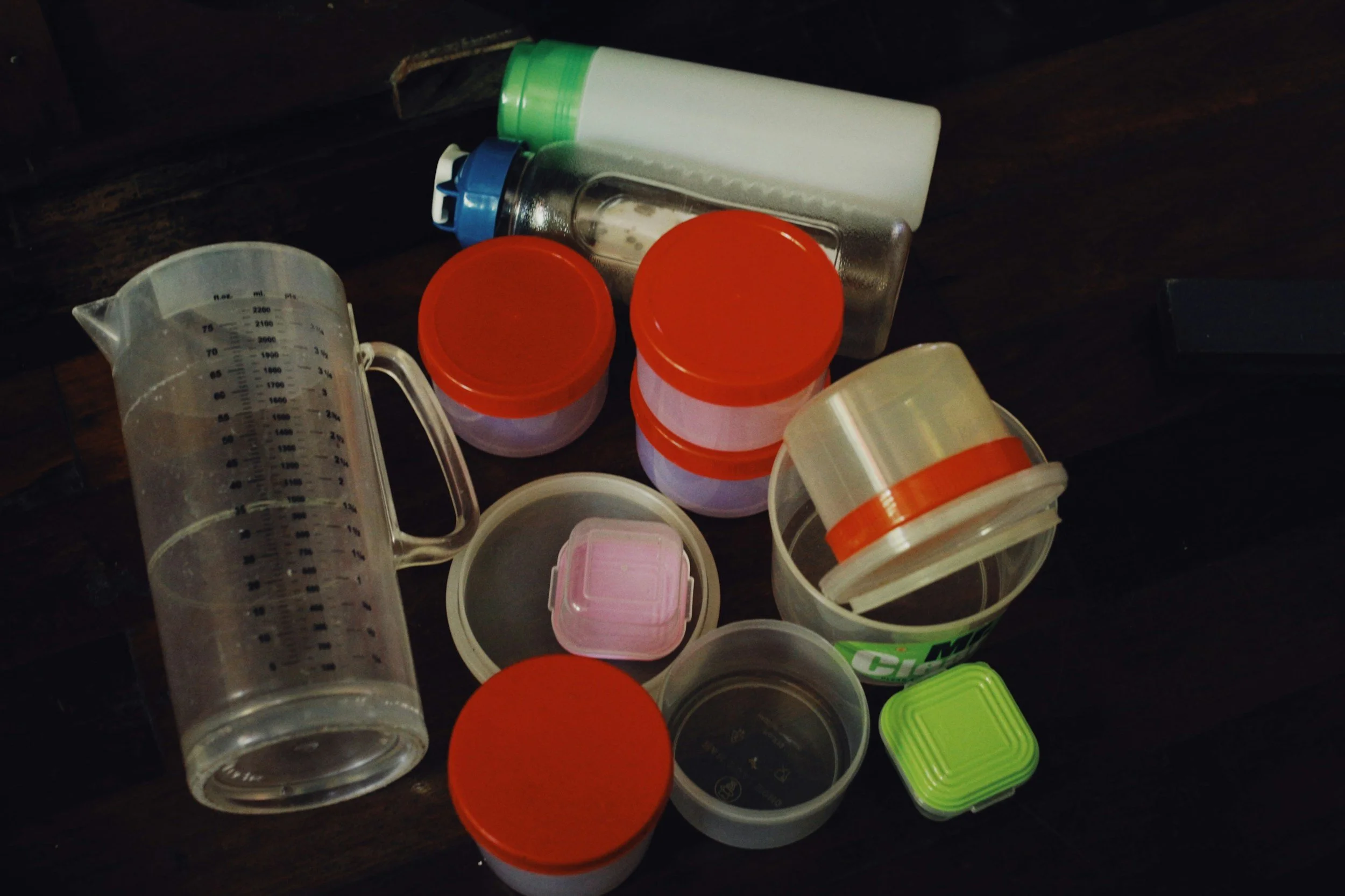 Collection of plastic containers and jars with colorful lids, measuring jug, and water bottles placed on a dark wooden surface.