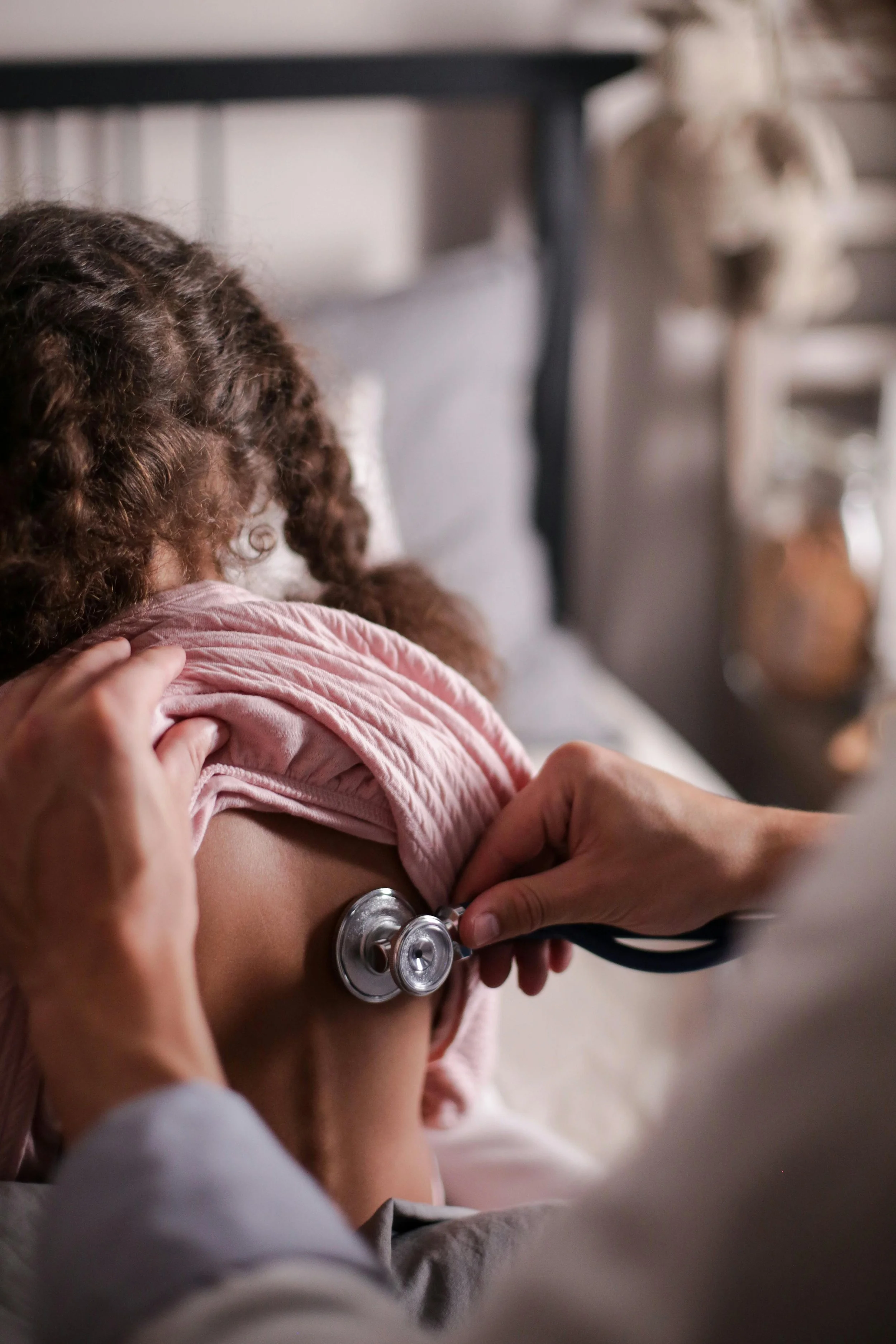 A healthcare professional using a stethoscope to examine a girl with curly hair in a pink shirt, in a medical setting.