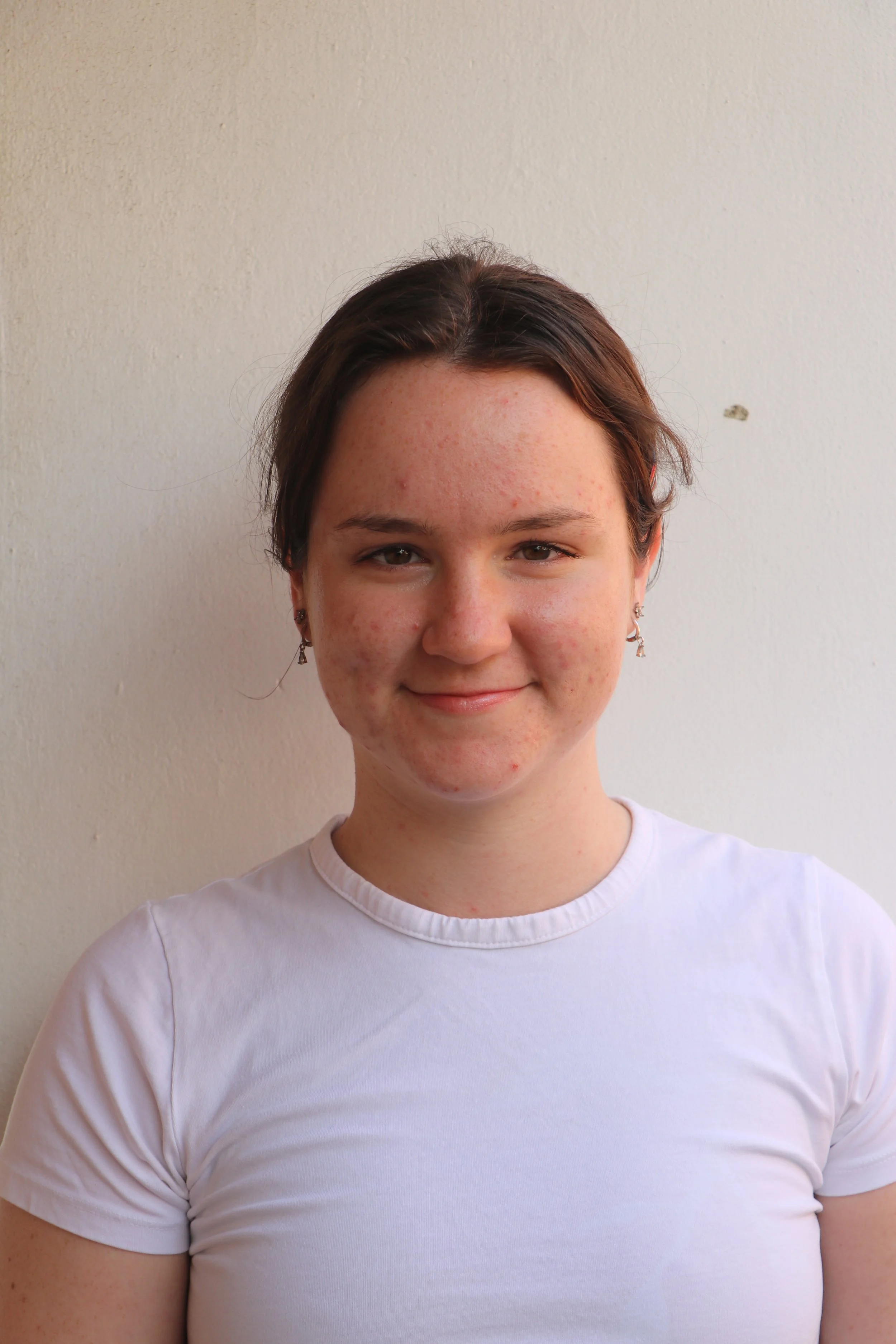 A young woman with short brown hair and earrings, wearing a white t-shirt, standing in front of a plain wall, smiling softly at the camera.