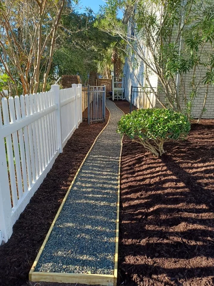 A narrow garden pathway with gravel, bordered by wooden edges, winding between a white picket fence and a house, with trees and shrubs casting shadows.