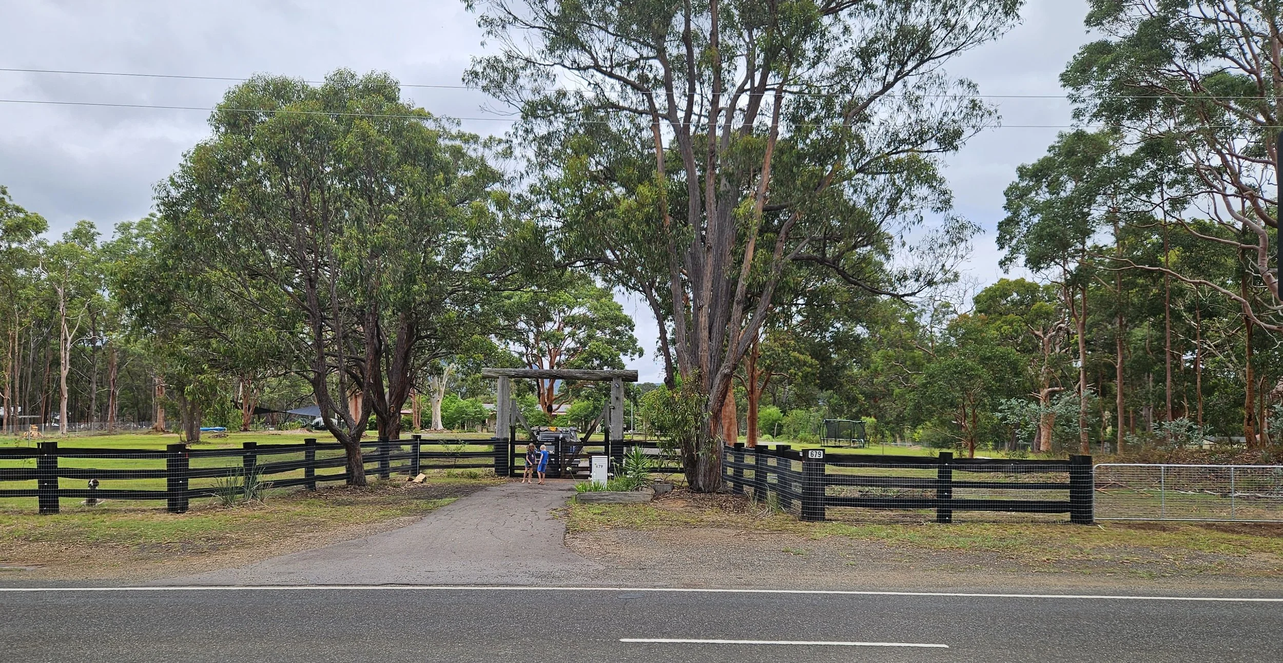 A rural scene with a gravel driveway leading to a fenced property with large trees and a wooden gate. The fence is black with horizontal slats, and there are two children standing near the gate. The background features more trees and a cloudy sky, with a divided road fronting the scene.