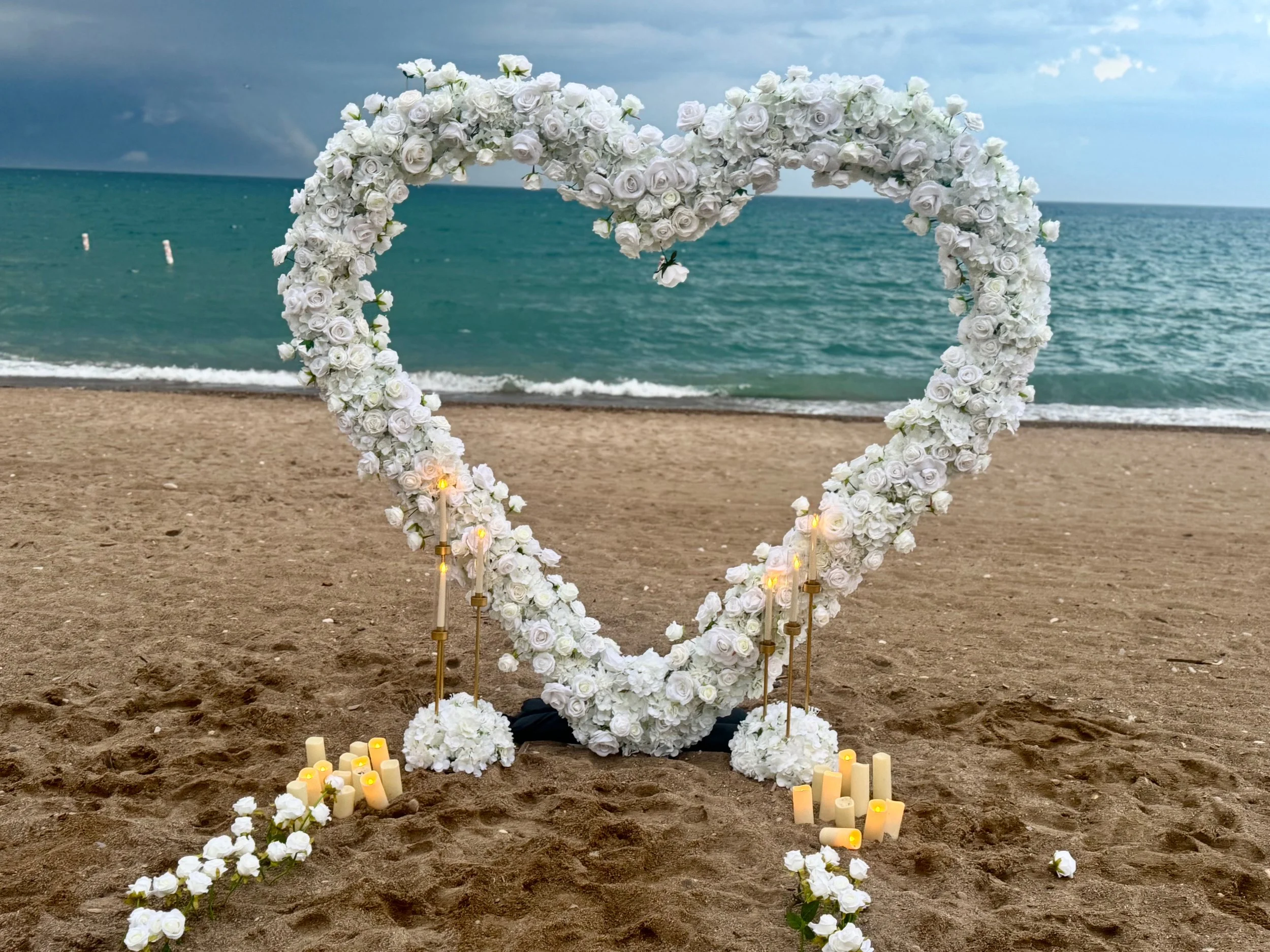 Heart-shaped floral arch made of white roses on a beach, with candles and candles holders at its base, overlooking the ocean during daytime.