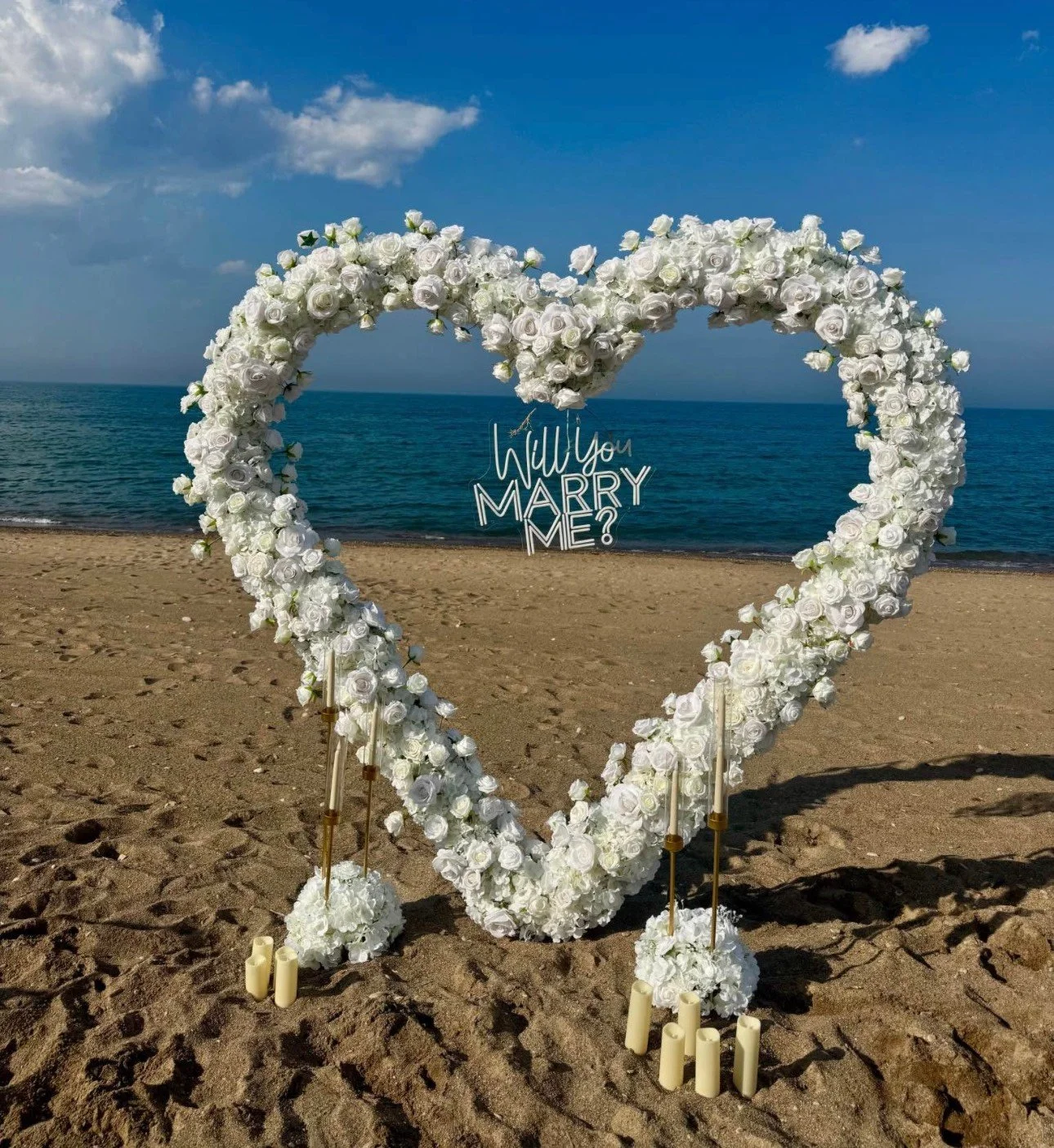 A heart-shaped floral arrangement made of white roses on a beach, with a neon sign asking 'Will you marry me?' inside the heart, candles at the base, and the ocean and sky in the background.