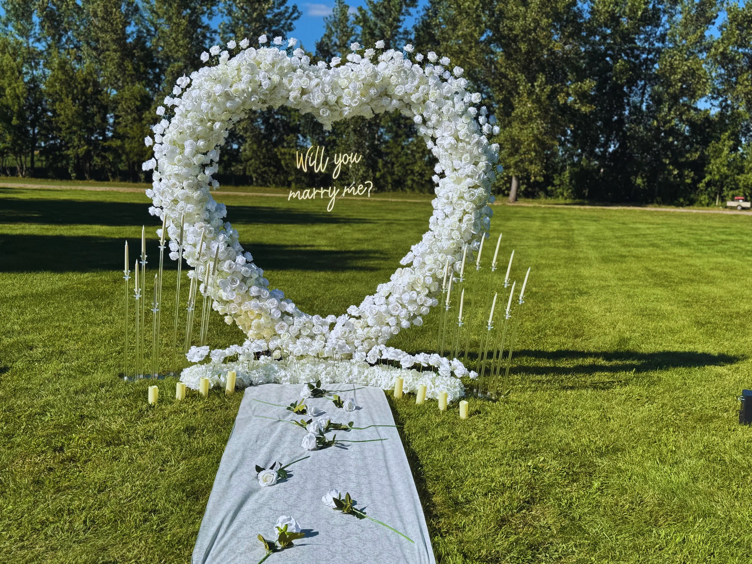 White rose flower arch with the words 'Will you marry me?' inside, set in a grassy field with trees in the background, candles and candles holders around the arch, and a white cloth with scattered roses on the ground.