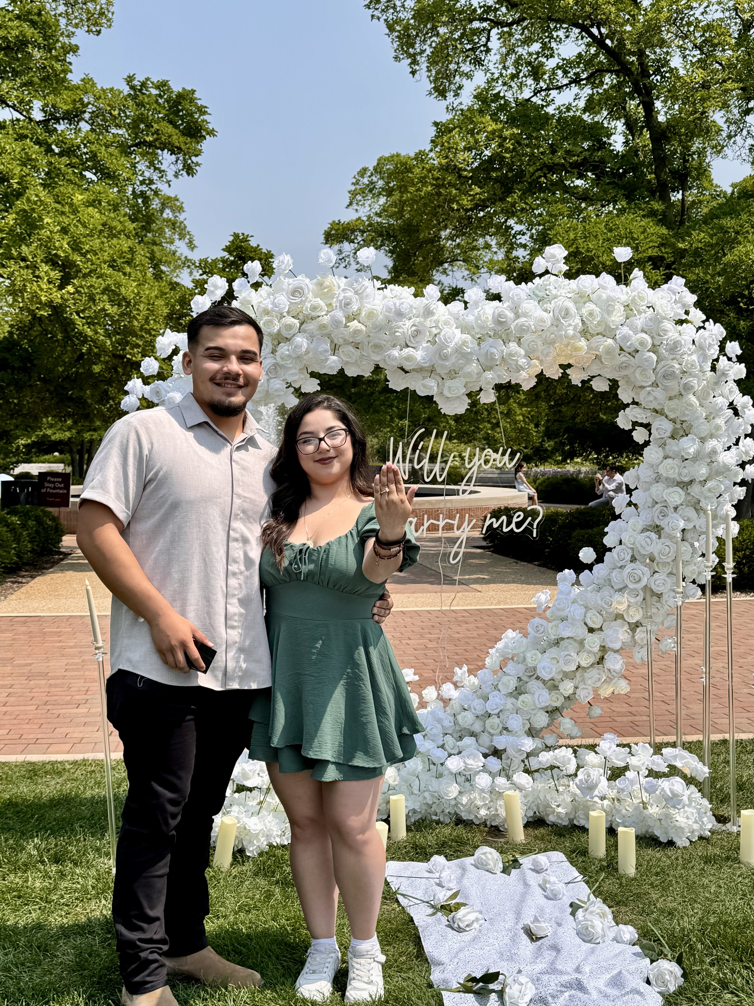 A couple stands in front of a large heart-shaped floral decoration with the words 'Will you marry me?' during a proposal at a park on a sunny day. The woman shows her engagement ring.