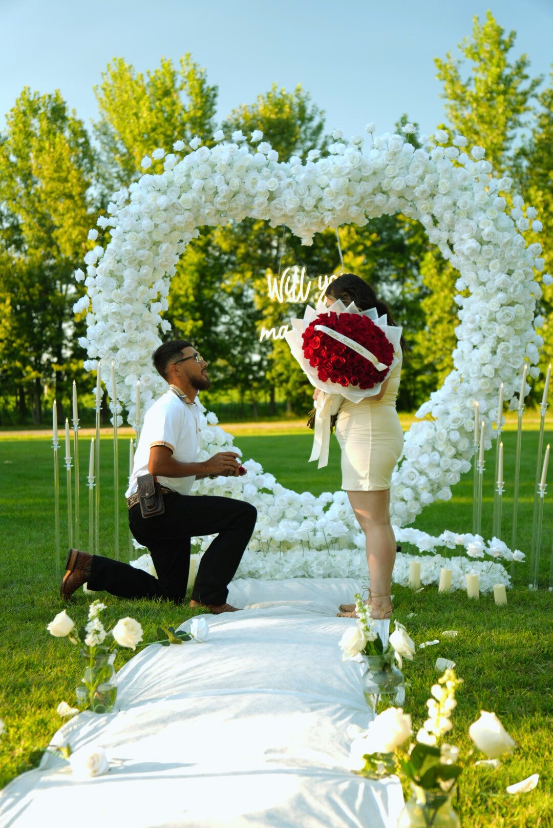 A man proposing to a woman outdoors during sunset, with a large heart-shaped floral arch decorated with white roses in the background, and candles on the grass.