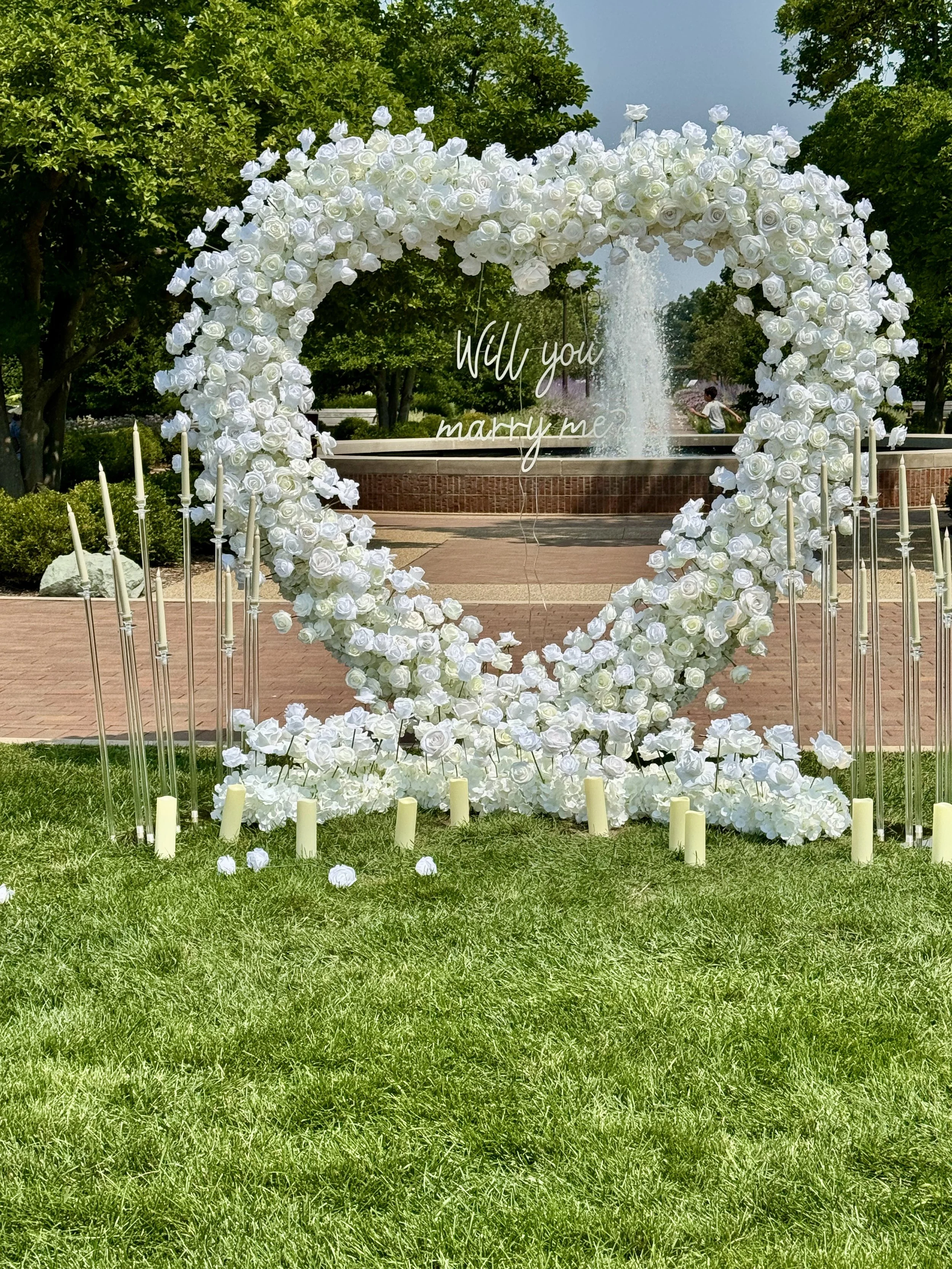 A white heart-shaped floral arch decorated with white roses, set outdoors near a fountain and trees, with candles at its base. A sign in the arch reads 'Will you marry me?'