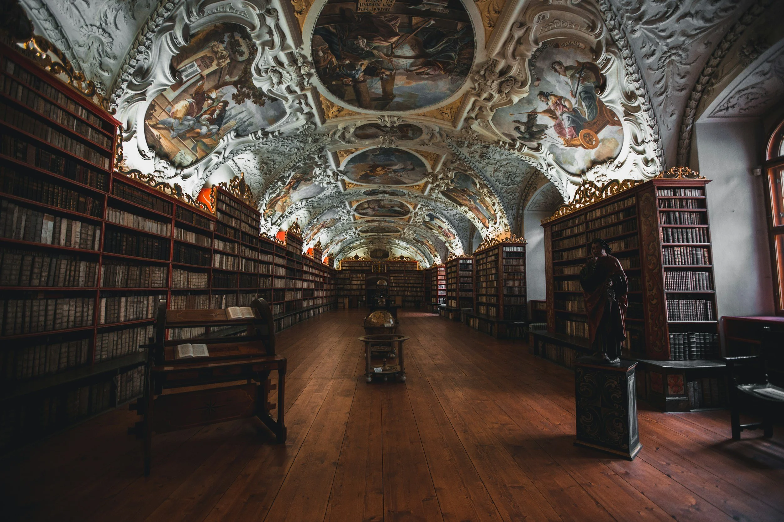 A grand, historic library with arched, painted ceilings, tall wooden bookshelves lining both sides, and a wooden floor. There are open books on some tables and a globe in the center. A statue of a person stands near the right window.