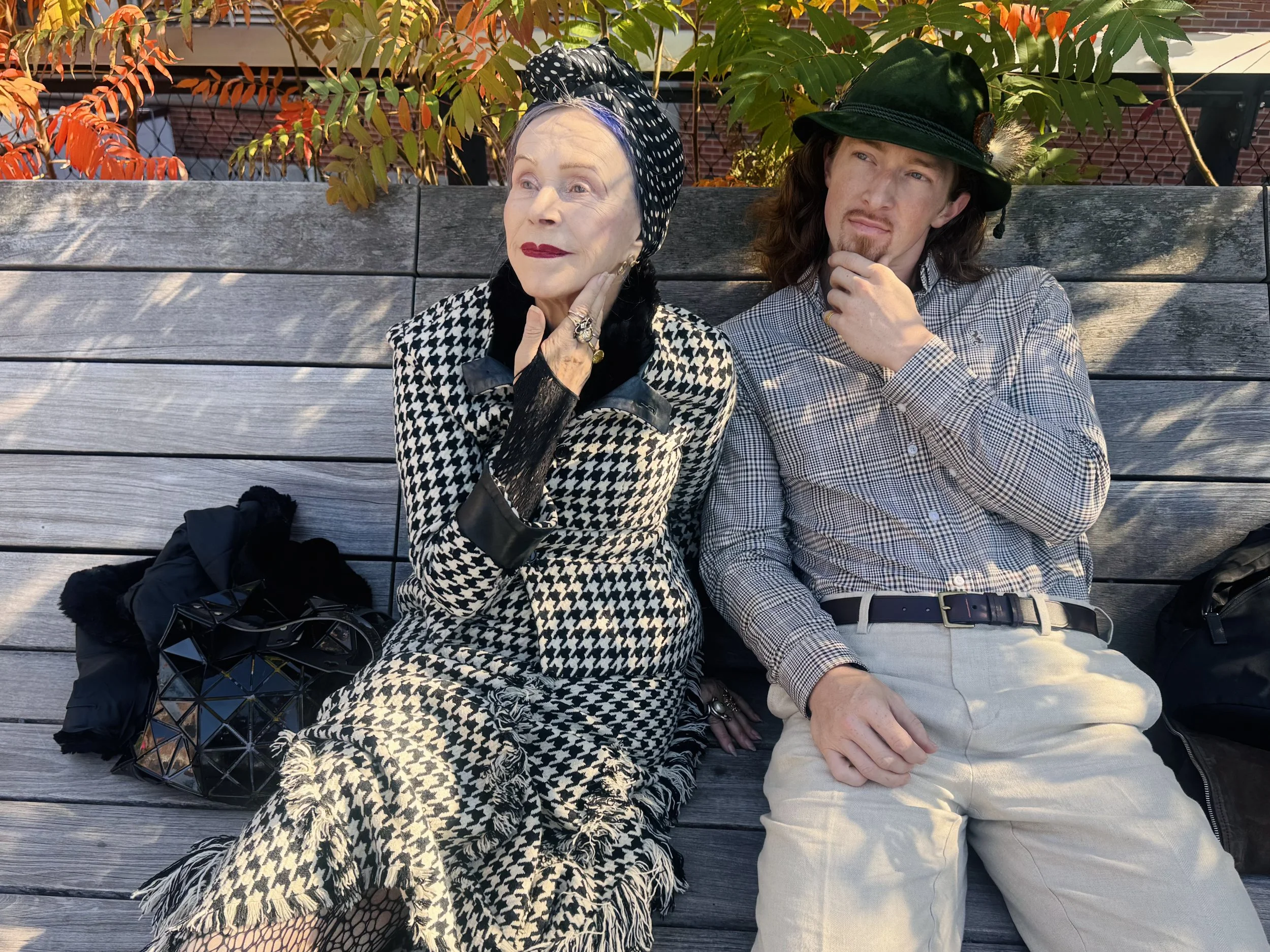 An elderly woman and a young man sitting on a wooden bench outdoors. The woman is wearing a black and white houndstooth patterned dress and headscarf, with her hand on her face, looking contemplative. The man is wearing a checkered shirt, beige pants, and a green hat, also appearing thoughtful, with his hand on his chin. There are bags and coat beside them, and autumn leaves are visible in the background.