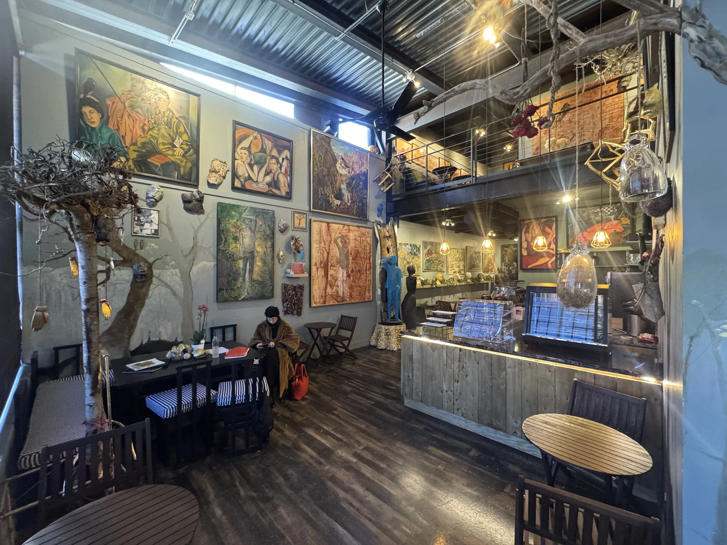 Interior of a cafe or art gallery with colorful paintings on the wall, decorative trees, hanging glass light fixtures, wooden furniture, and a person sitting at a table with a brown coat and red bag.