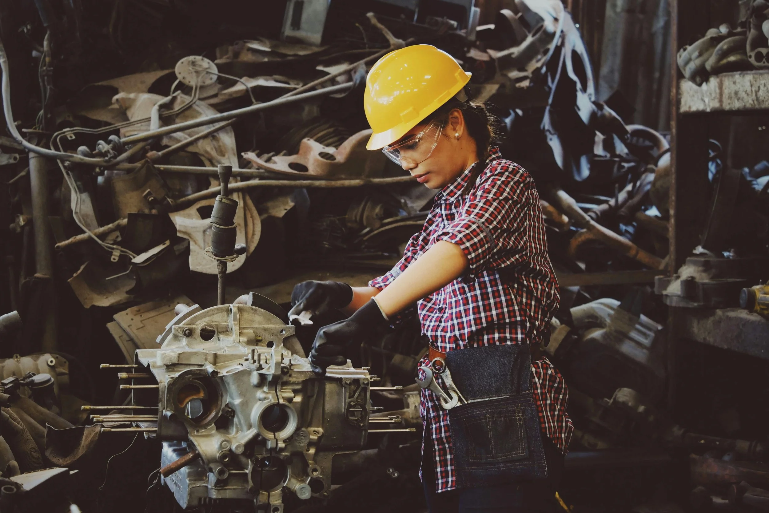 A woman wearing a yellow safety helmet, glasses, gloves, and a checkered shirt working on a car engine in a cluttered workshop.