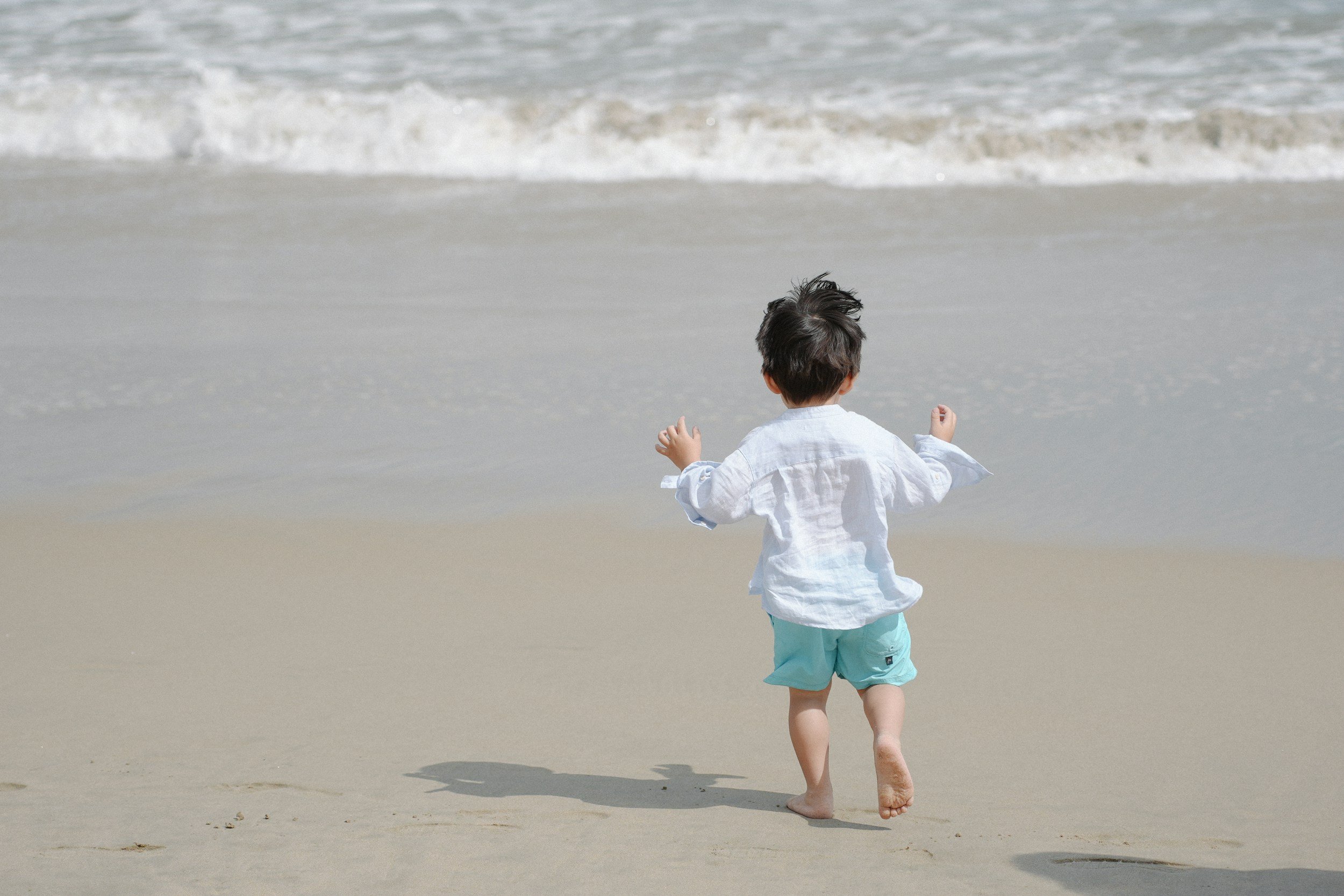 A young boy with dark hair wearing a white shirt and turquoise shorts walking barefoot on the sandy beach towards the ocean waves