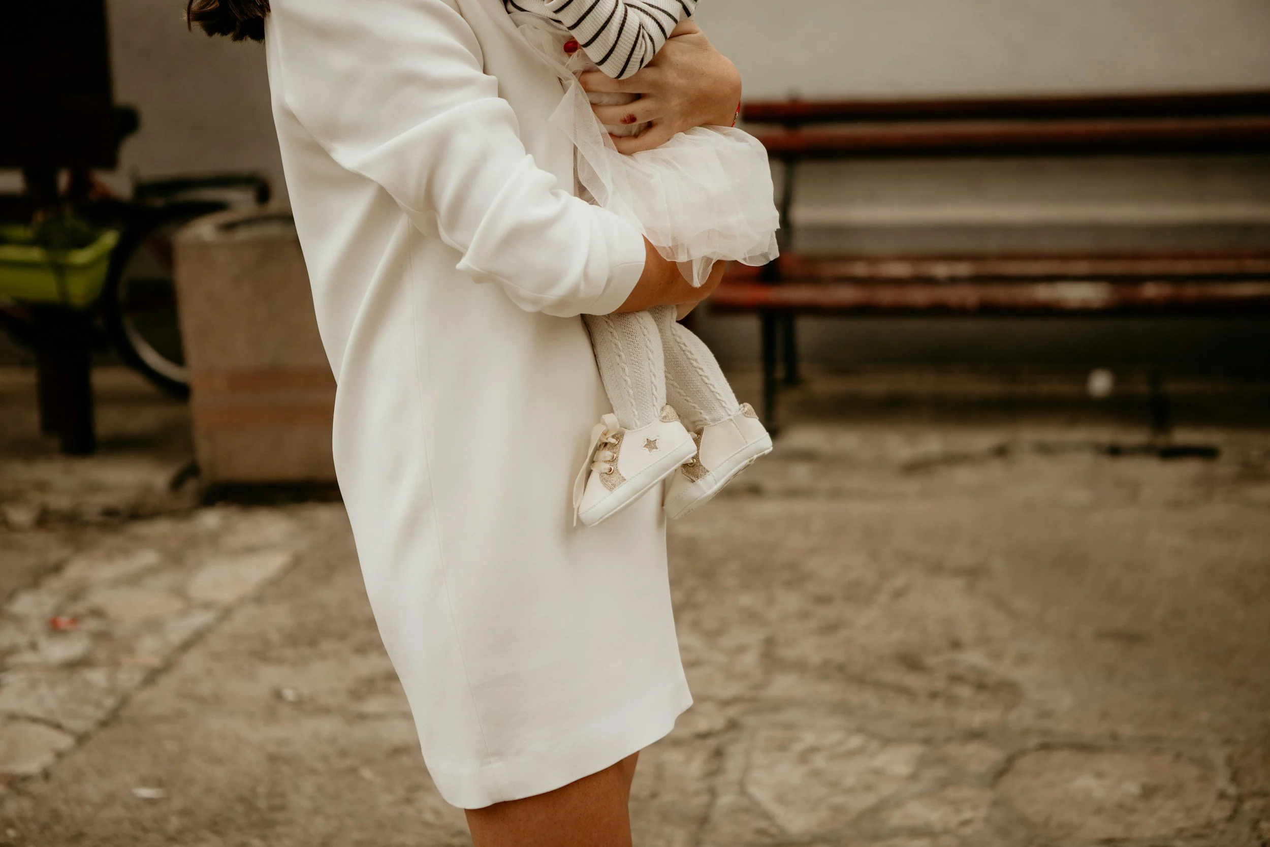 A woman holds a young child dressed in white shoes and a white dress. The woman is wearing a white coat and the child is dressed in a striped shirt under a white tulle dress, with the background featuring benches and a bicycle.
