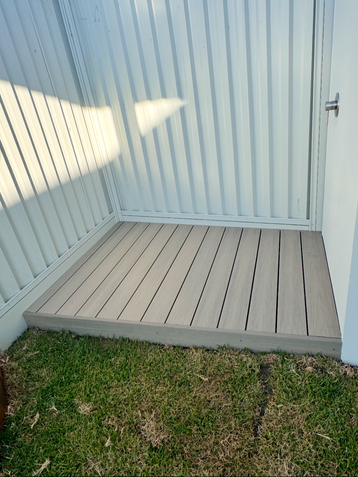 Small outdoor deck with wooden planks, adjacent to a patch of grass, enclosed by light-colored metal fencing and a white wall.
